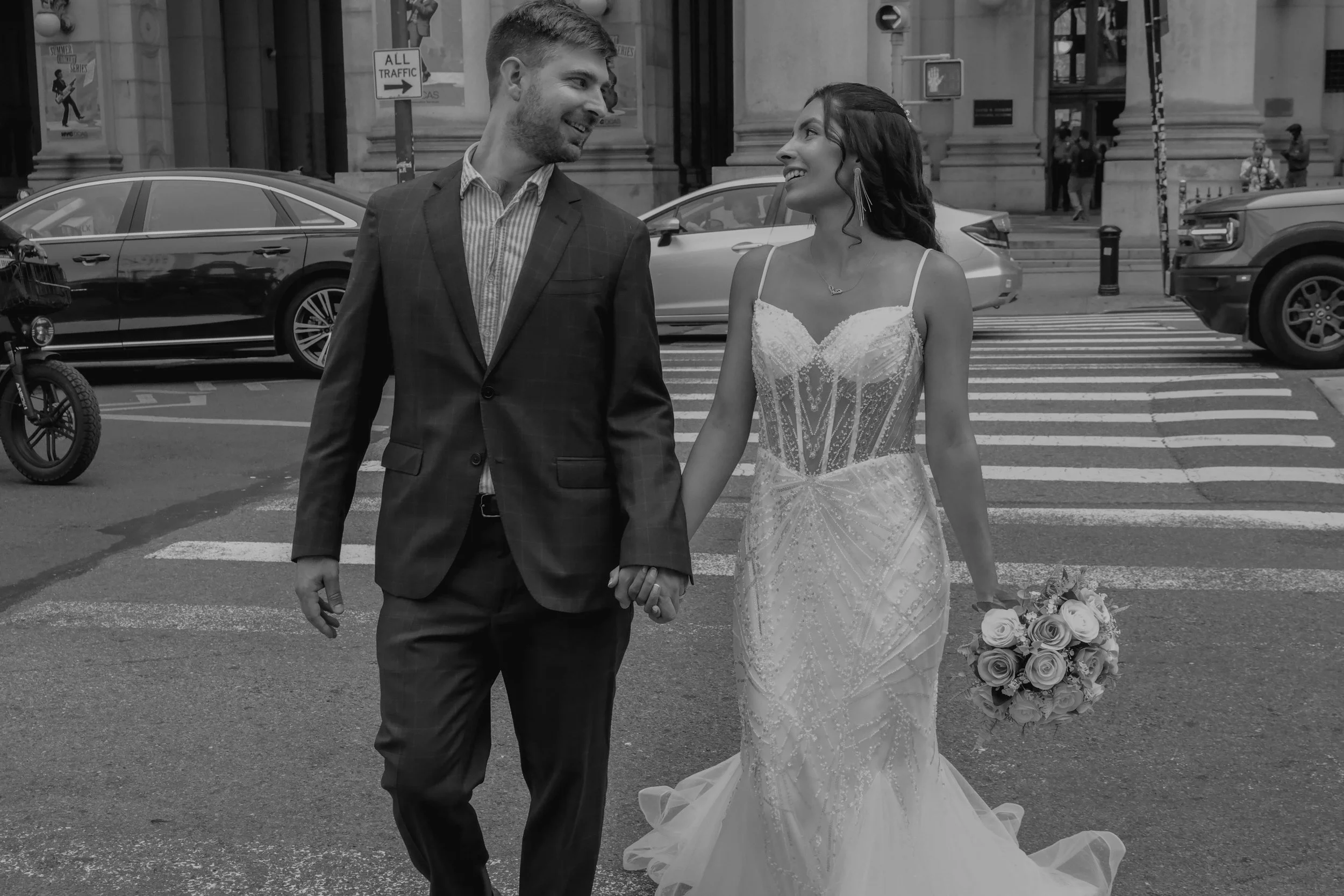 A couple in wedding attire holding hands and smiling at each other while walking across a city street.