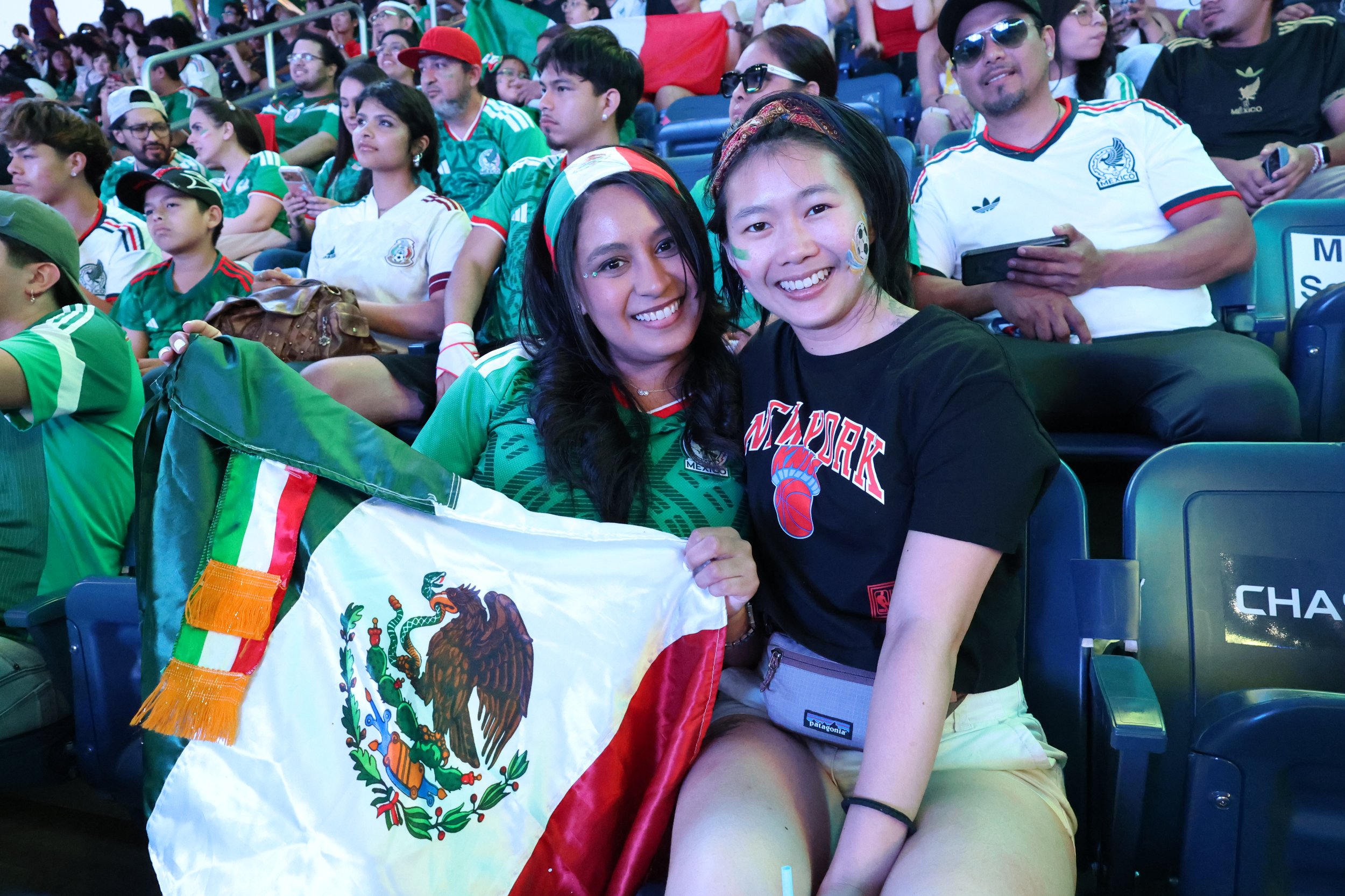 A group of people, mostly wearing Mexican national soccer team jerseys, sitting in stadium seats at a sports event. Two women in the foreground are smiling and holding a Mexican flag, celebrating together.