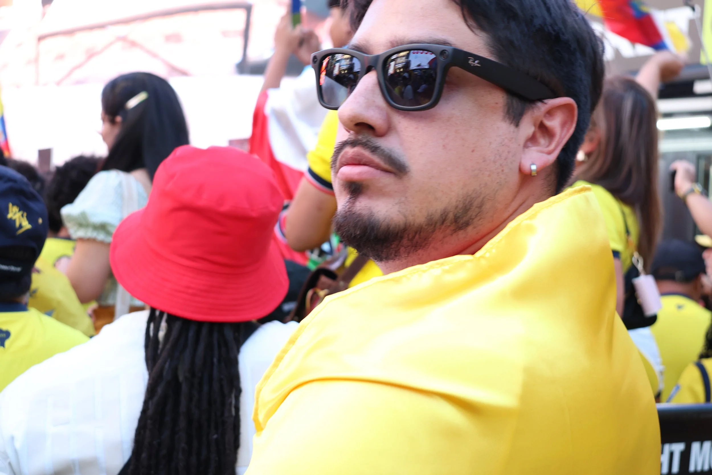 Close-up of a man wearing black sunglasses and a yellow jacket, surrounded by a crowd of people wearing colorful clothing at an outdoor event.