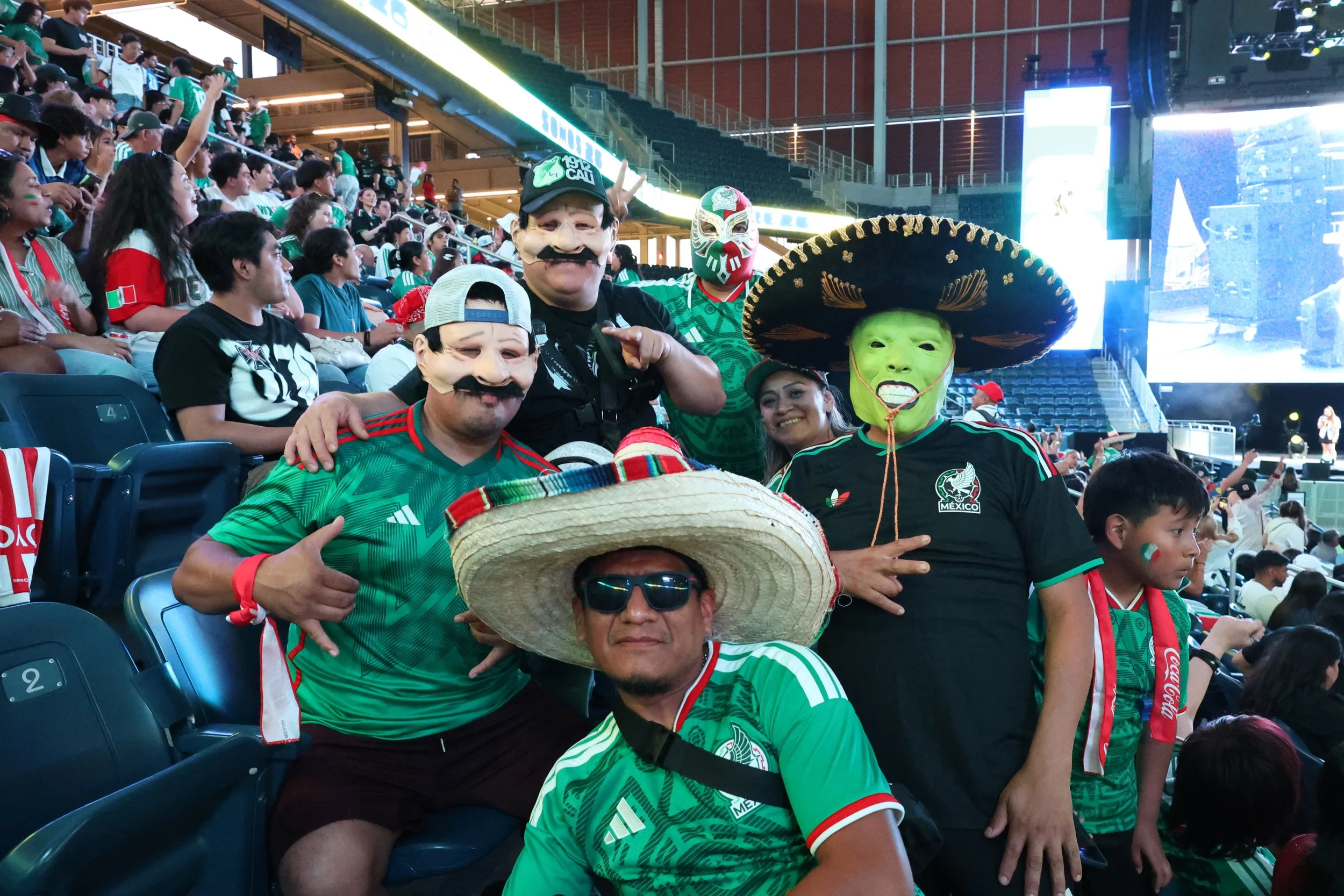 Group of soccer fans wearing green jerseys and Mexican masks at a stadium, celebrating during a match.