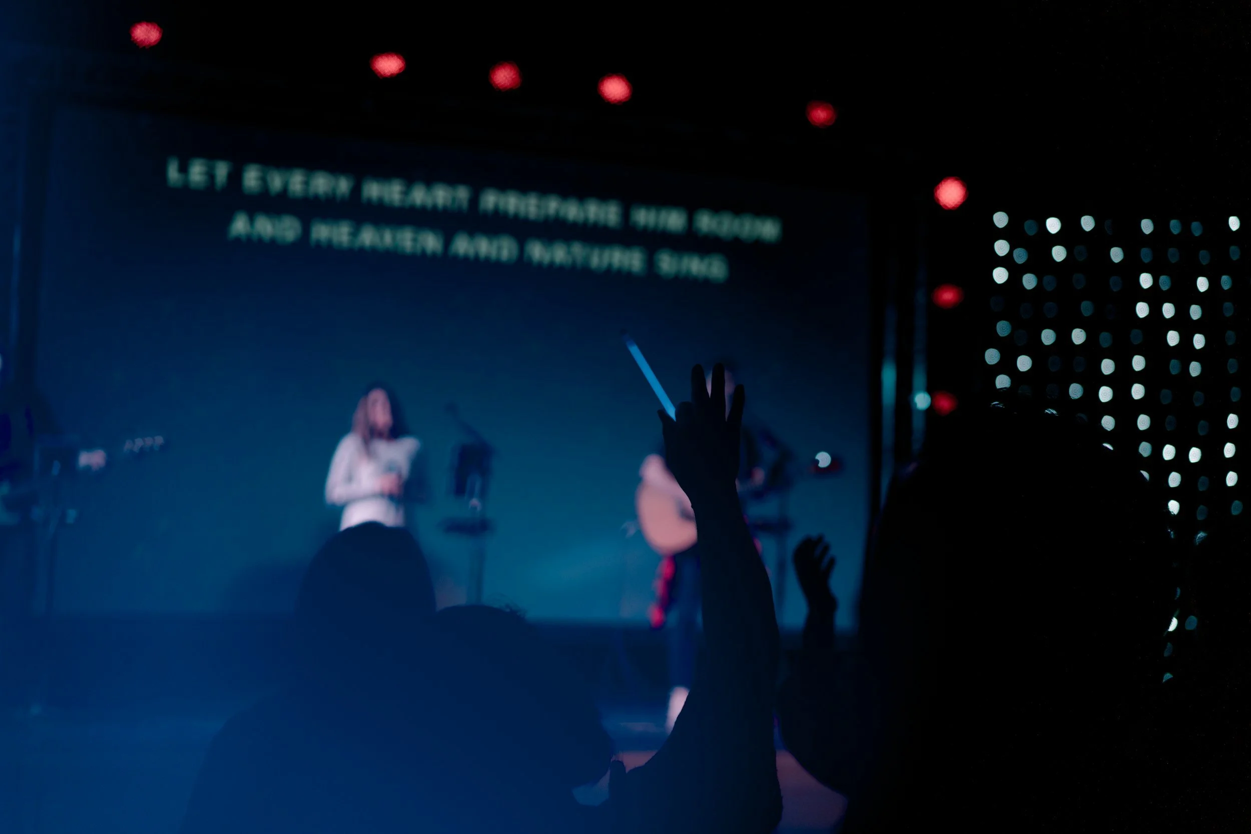 Concert scene with a stage, a singer with a guitar, and audience members raising hands, illuminated by colorful lights.