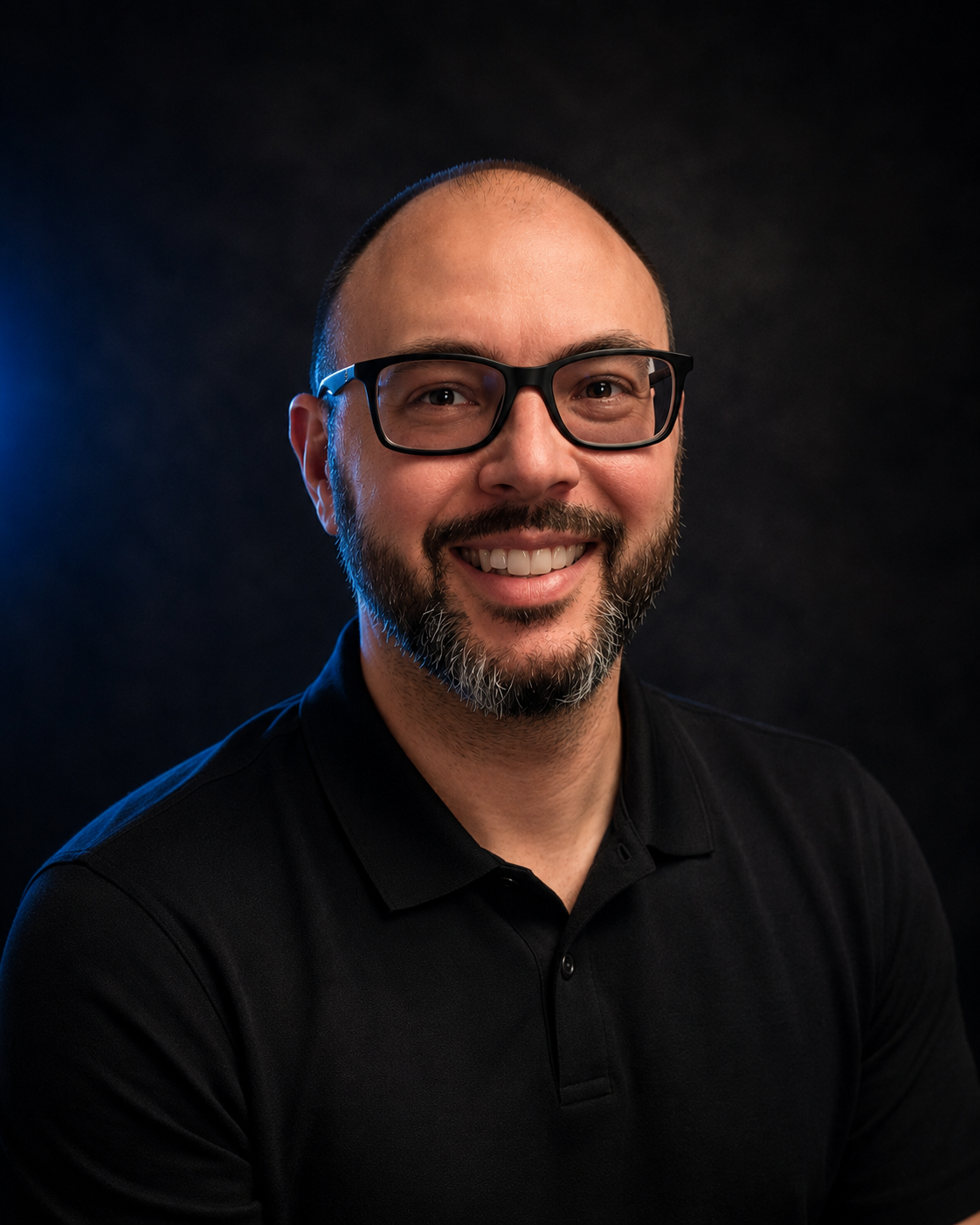 Smiling man with glasses, beard, and mustache wearing a black polo shirt against a dark background.