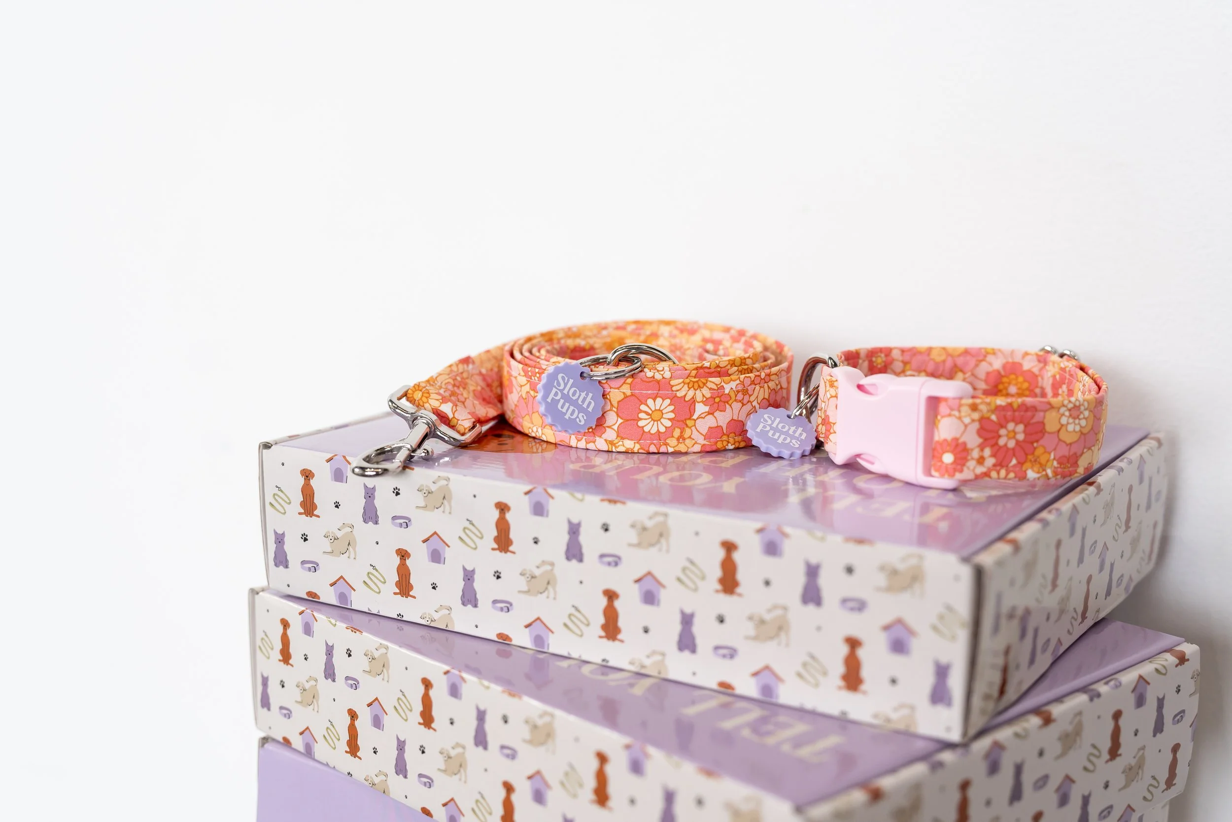 Dog collars decorated with a pink floral pattern, placed on top of stacked decorated boxes, some with illustrations of dogs and houses.