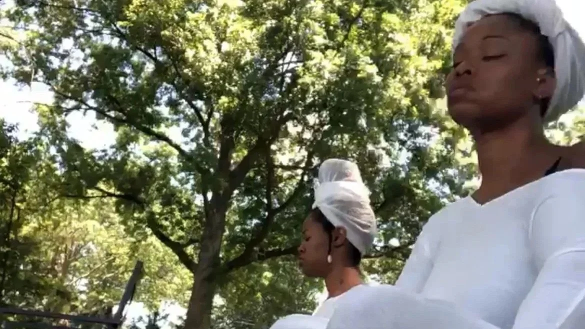 Two women dressed in white with head wraps, sitting outdoors under a tree, appearing to meditate or practice mindfulness.