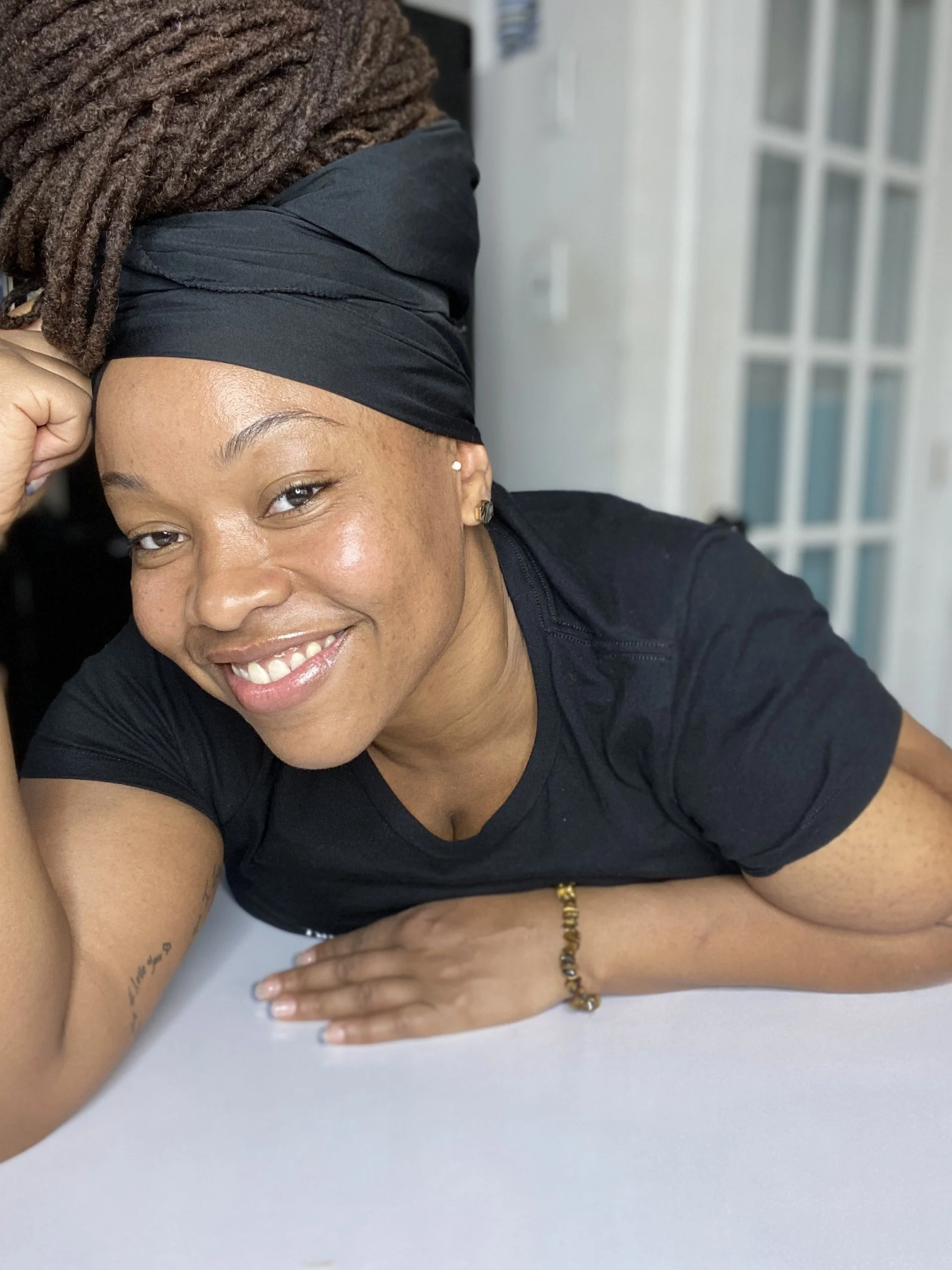 Close-up of a smiling woman with locs, wearing a black headscarf and black shirt, sitting at a white table with her left arm resting on the surface and a gold bracelet on her wrist.
