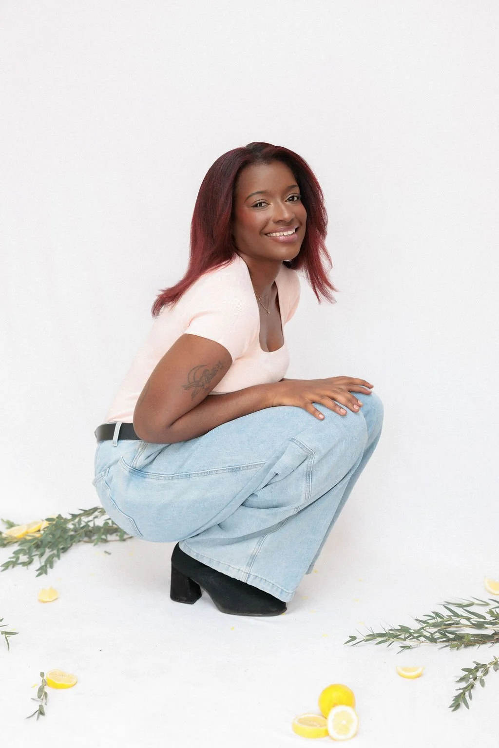 A woman with red hair squatting, smiling, wearing a light pink shirt, high-waisted jeans, and black shoes, surrounded by lemon slices and green leafy branches on a white background.