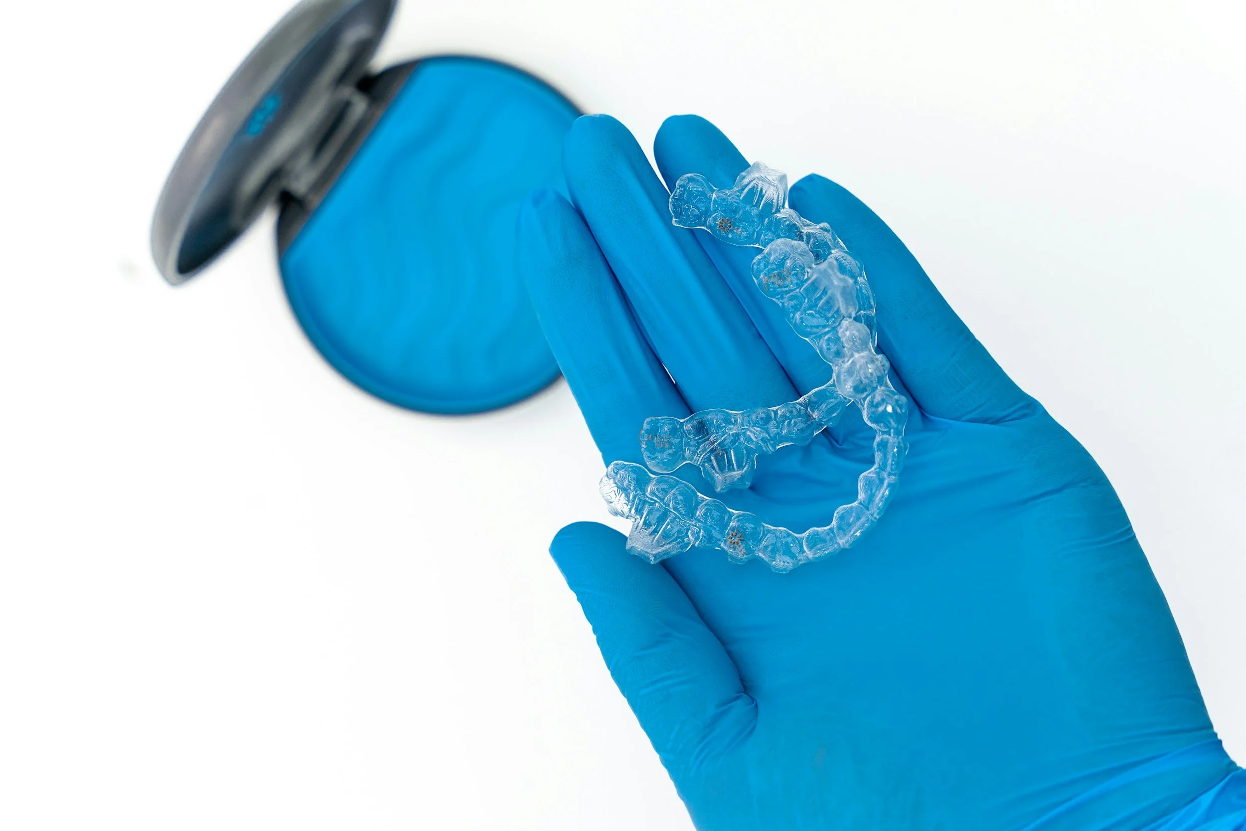 A gloved hand holding a clear dental aligner, with a container in the background.