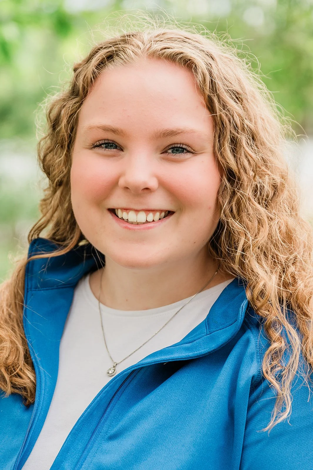 A smiling young woman with curly blonde hair, wearing a blue jacket and a necklace, outdoors with a blurred green background.