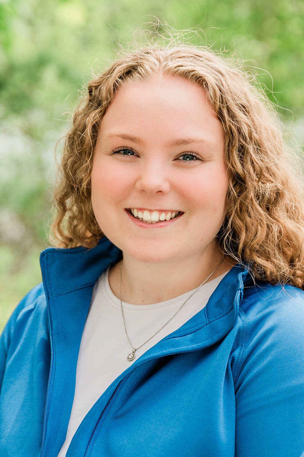 A smiling young woman with curly blonde hair, wearing a blue jacket and a necklace, outdoors with a blurred green background.