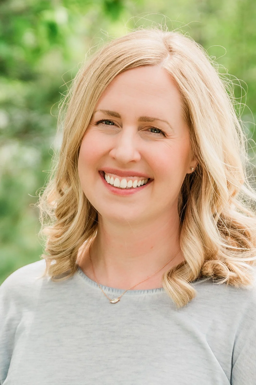 Dr. Bree Highstrom, dentist at Legacy Family Dental in Eagle River, WI. A woman with blonde hair, smiling, wearing a light gray top and a delicate necklace, outdoors with green foliage in the background.