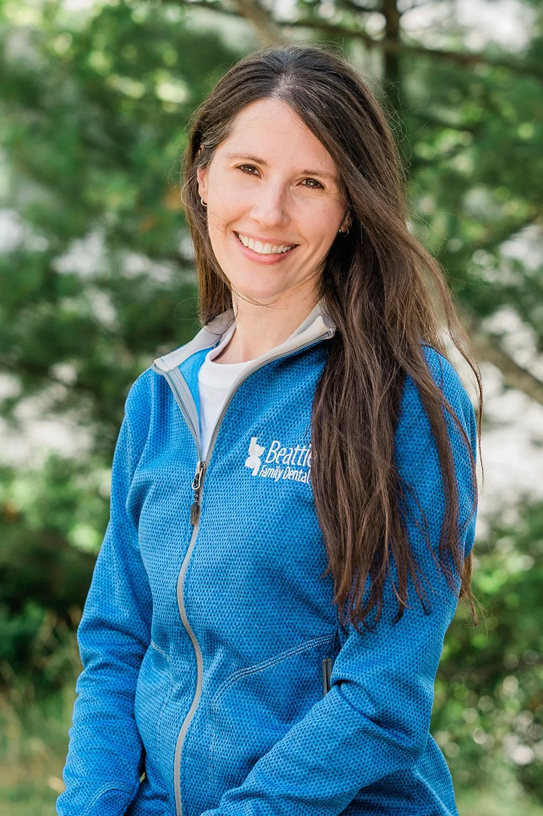 A smiling woman with long brown hair, wearing a blue jacket with the logo 'Beattie Family Dentistry,' standing outdoors with green trees in the background.