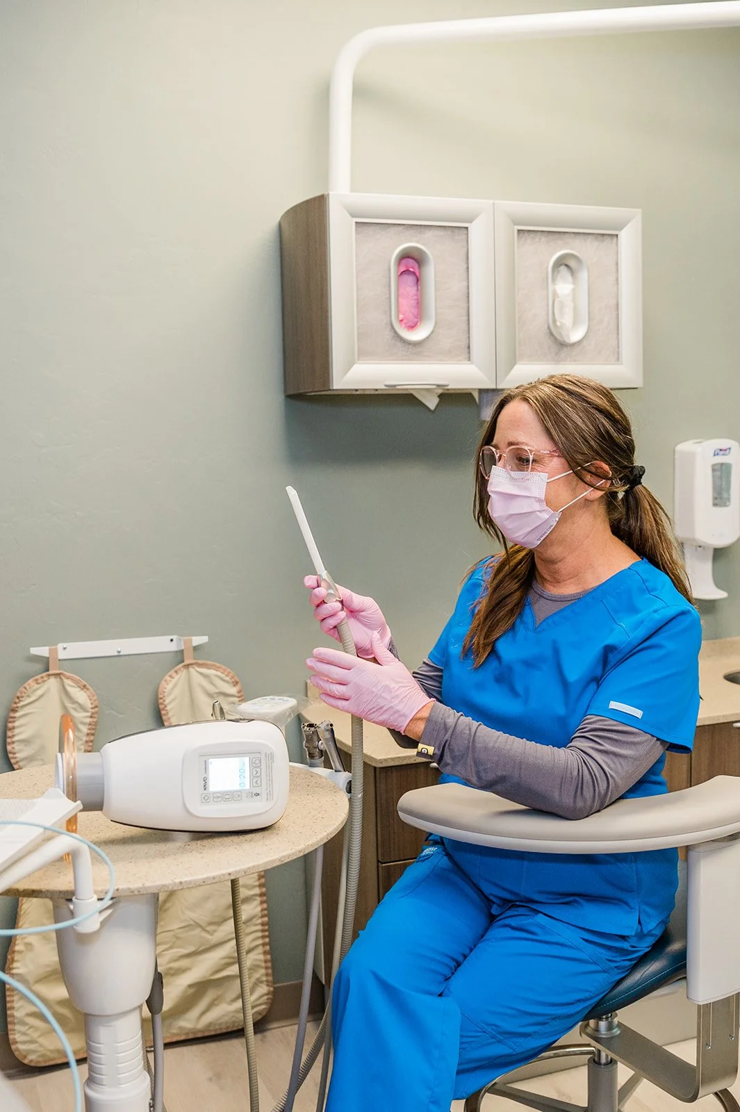 A healthcare worker in blue scrubs and a pink face mask sitting in the Legacy Family Dental office. She is holding a dental instrument, with dental gloves on, in front of her, surrounded by medical equipment and supplies.