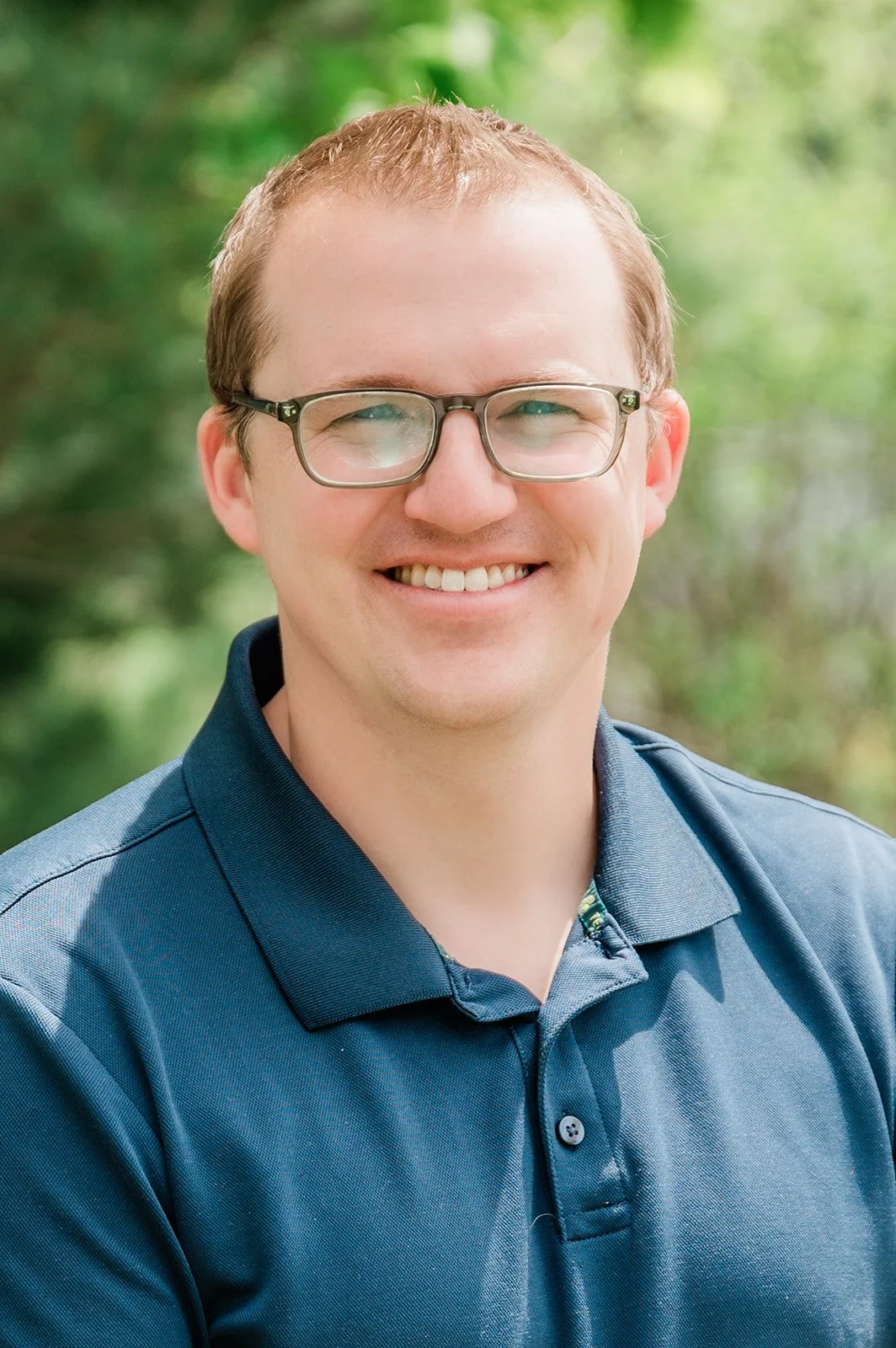 Dr. Brad Camp, owner of Legacy Family Dental, and dentist at the practice. Photo of a smiling man with glasses, wearing a dark blue polo shirt, outdoors with blurred green trees in the background.