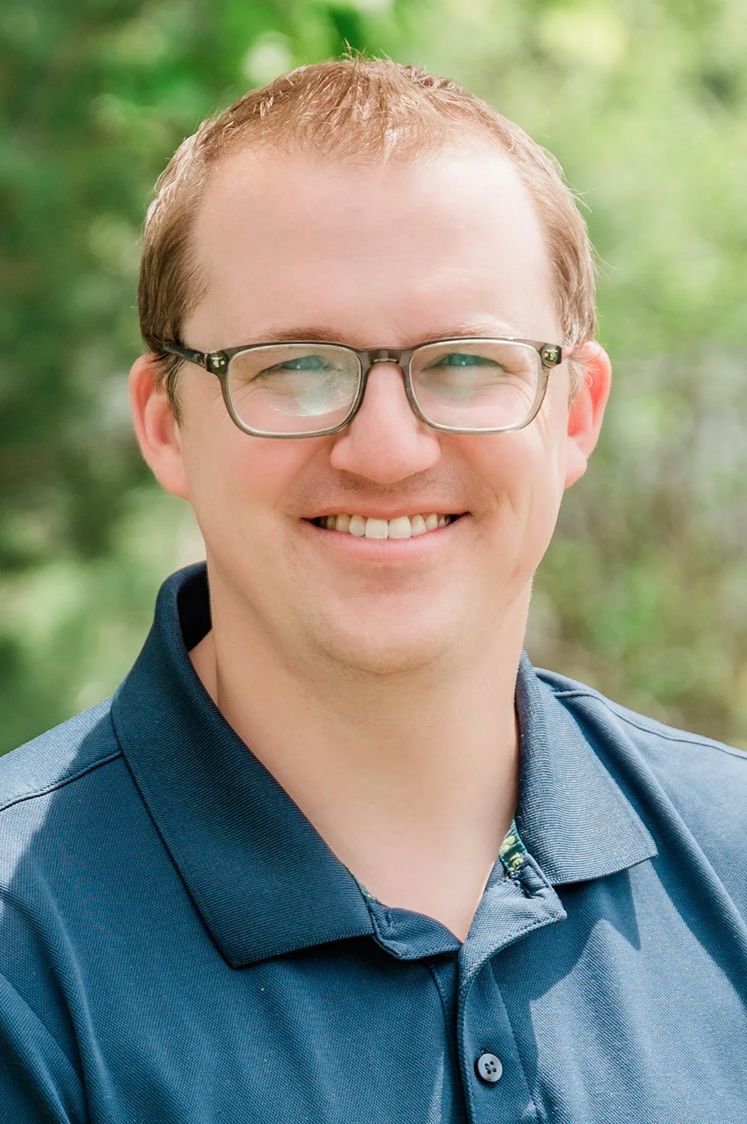 Dr. Brad Camp, owner of Legacy Family Dental, and dentist at the practice. Photo of a smiling man with glasses, wearing a dark blue polo shirt, outdoors with blurred green trees in the background.