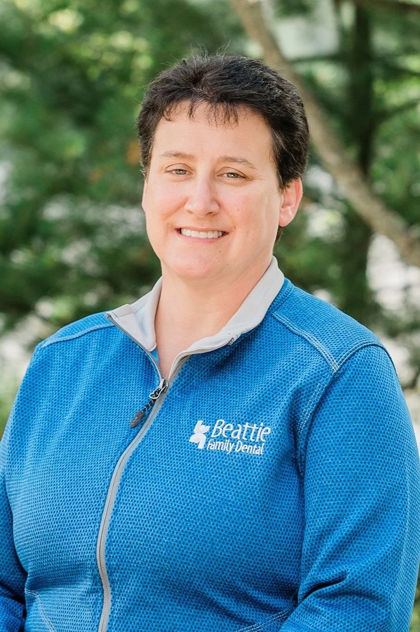 A woman smiling outdoors wearing a blue jacket with 'Legacy Family Dental' logo, with trees in the background. This is Roni, a dental hygienist.
