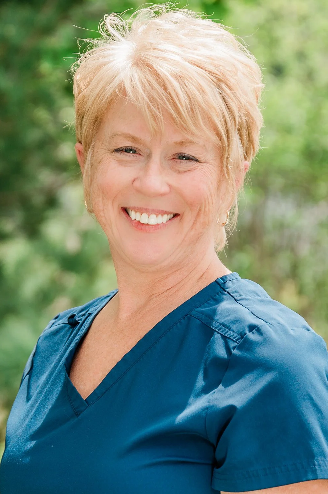 Portrait of Jacki (a dental hygienist at Legacy Family Dental), a smiling middle-aged woman with short blonde hair, wearing blue scrubs, outdoors with blurred green foliage background.