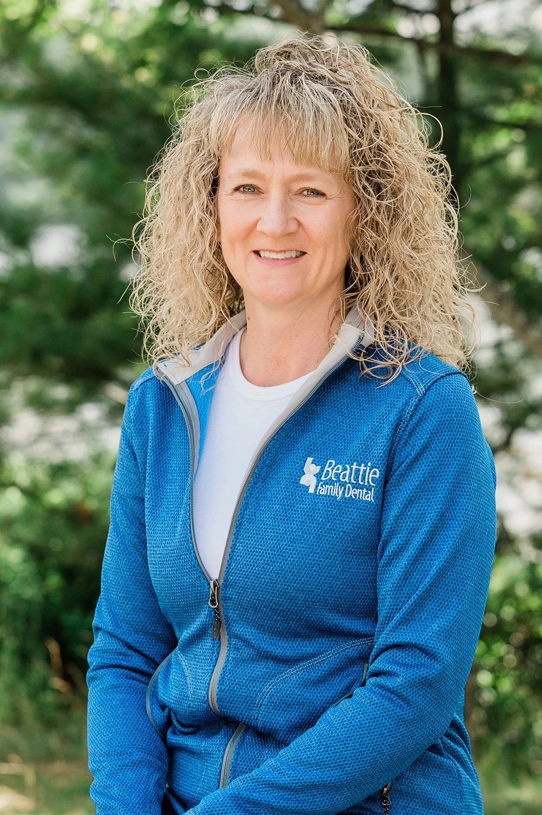 A woman with curly blonde hair smiling outdoors, wearing a blue zip-up jacket with 'Beattie Family Dental' logo.