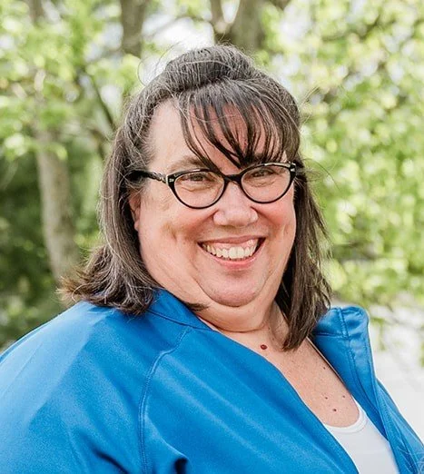 A person with long, brown hair and glasses smiling outdoors, wearing a blue jacket and a white shirt, with a blurred green background.