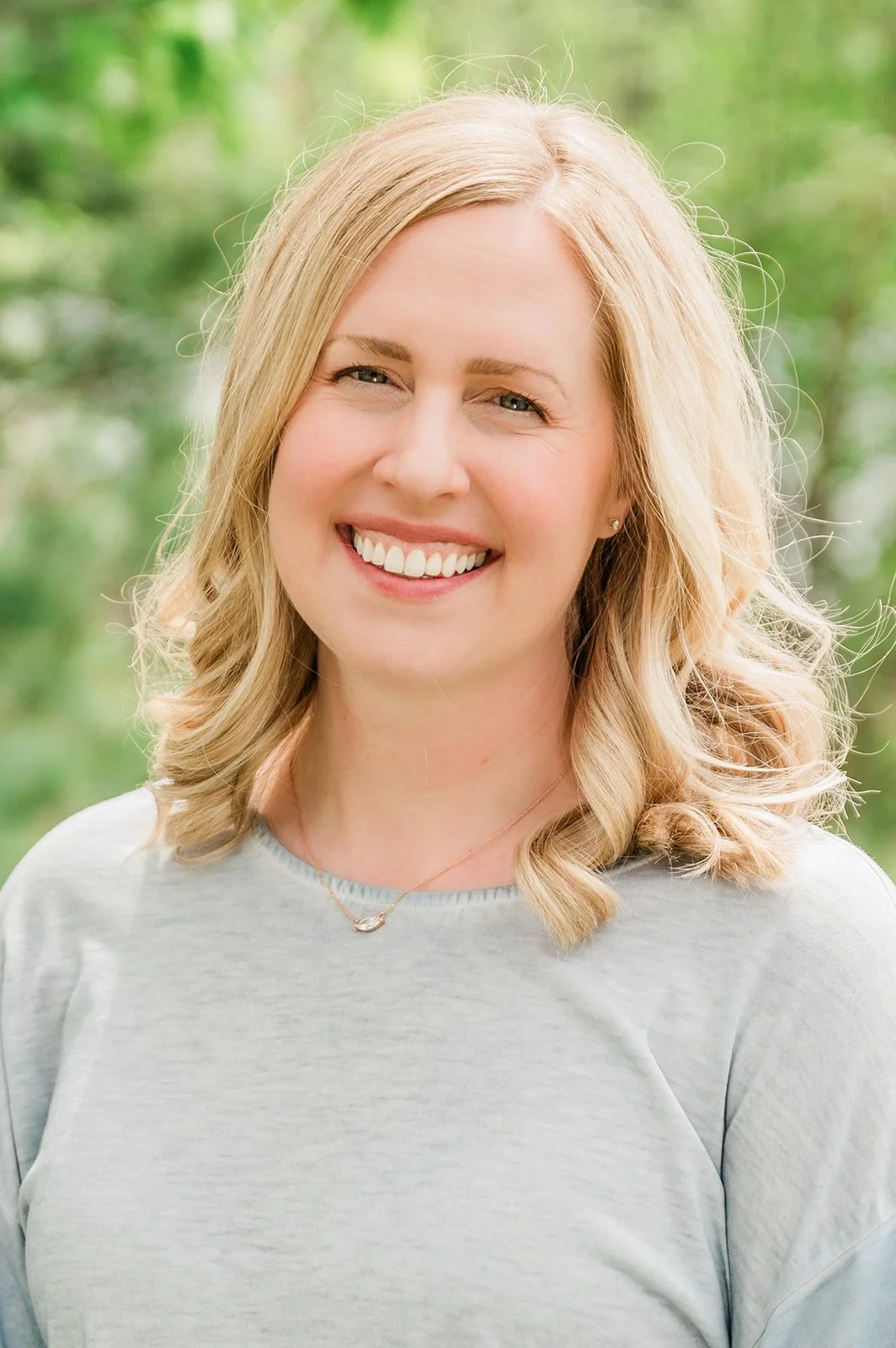 Dr. Bree Highstrom, dentist at Legacy Family Dental in Eagle River, WI. A woman with blonde hair, smiling, wearing a light gray top and a delicate necklace, outdoors with green foliage in the background.