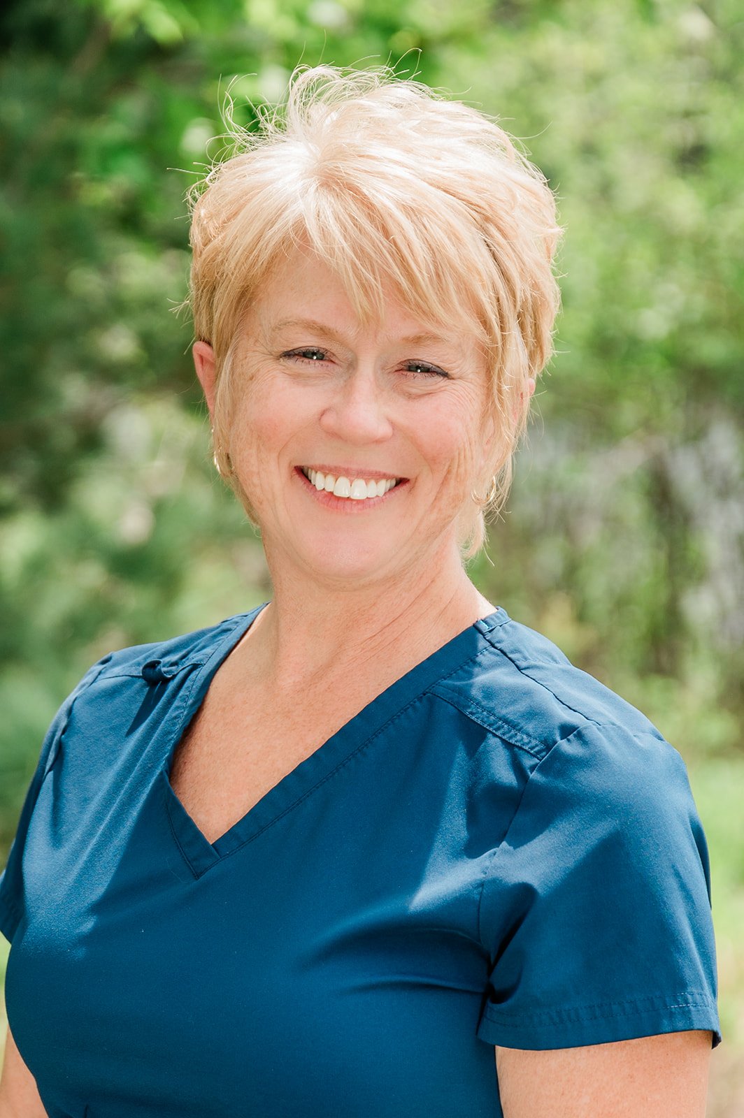 Portrait of Jacki (a dental hygienist at Legacy Family Dental), a smiling middle-aged woman with short blonde hair, wearing blue scrubs, outdoors with blurred green foliage background.