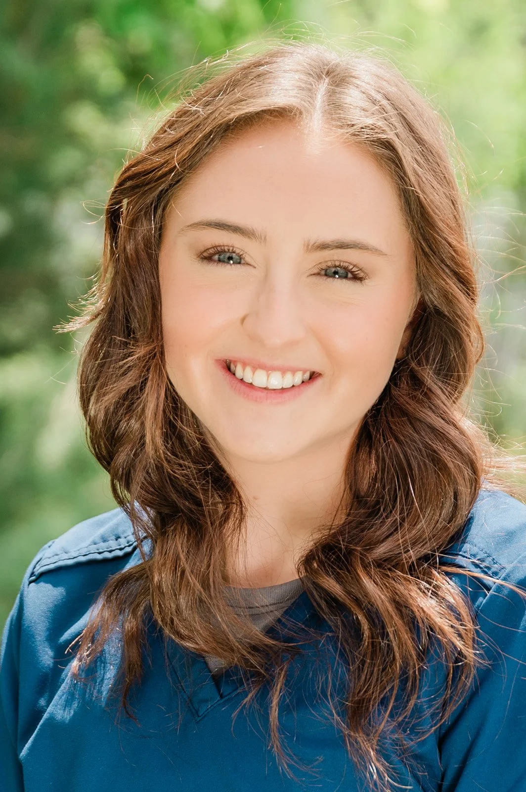 A woman with wavy brown hair, blue eyes, and a bright smile, wearing a blue jacket, outdoors with a blurred green background.