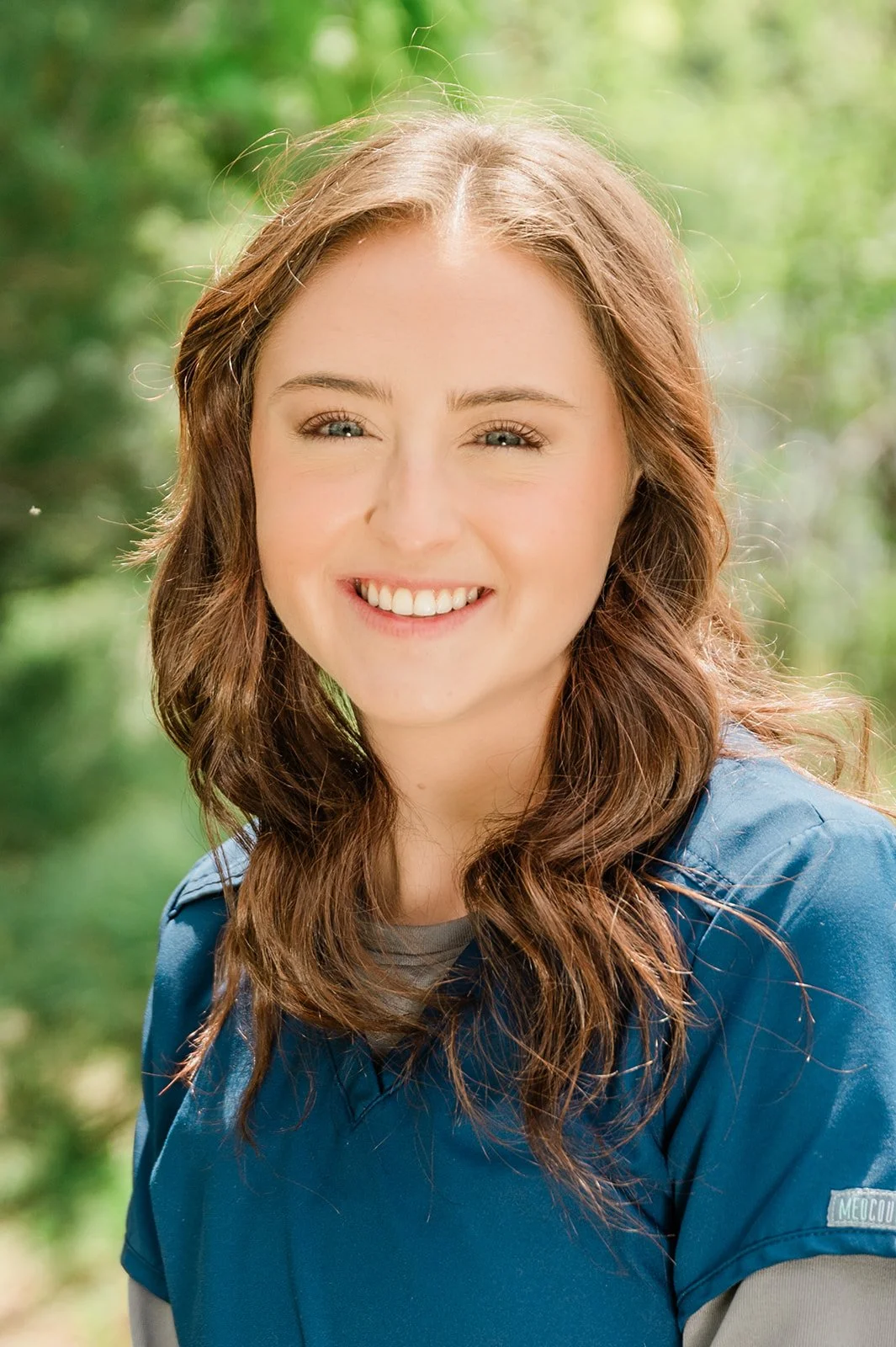 A woman with wavy brown hair, blue eyes, and a bright smile, wearing a blue jacket, outdoors with a blurred green background.