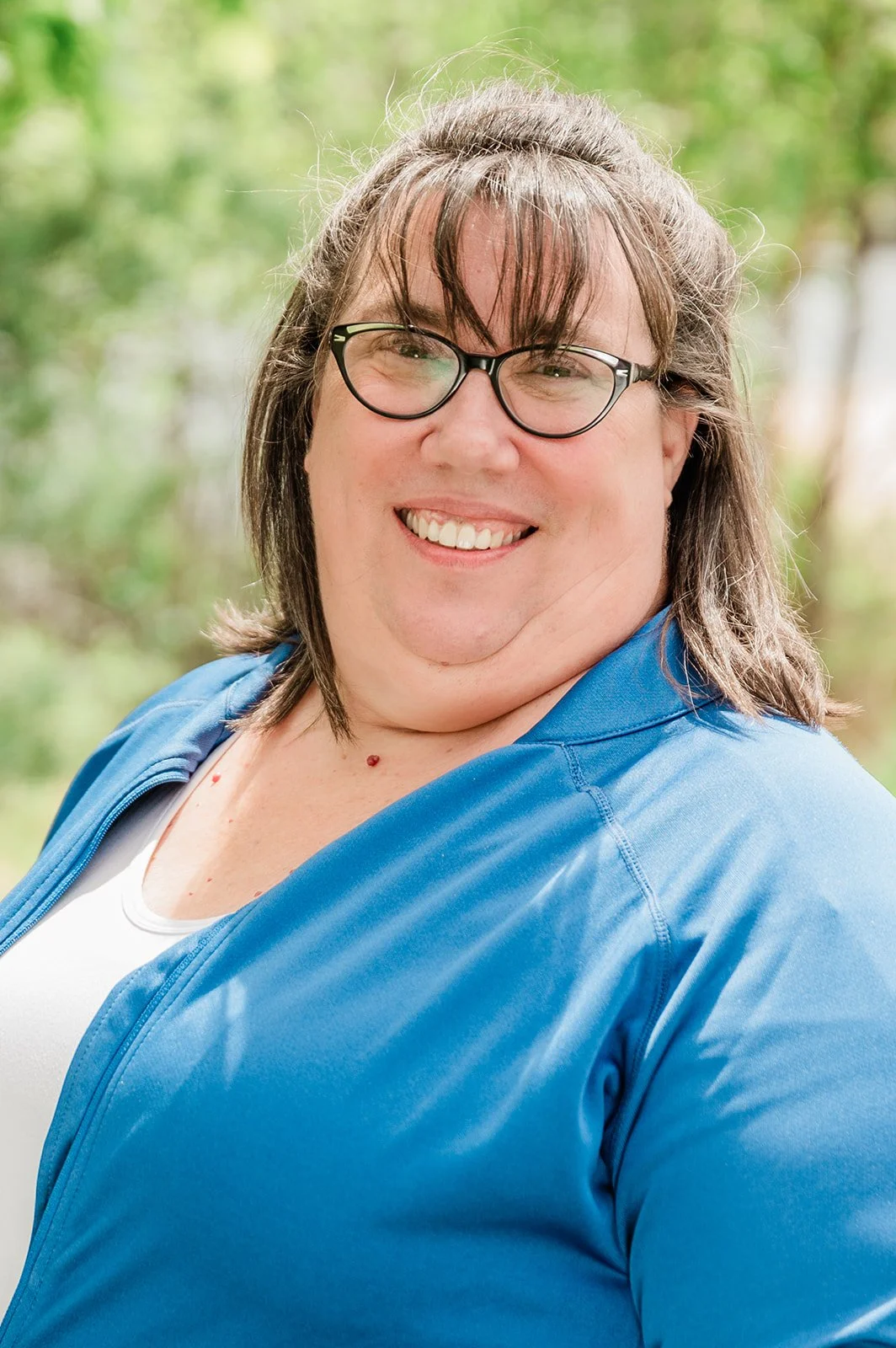 A person with long, brown hair and glasses smiling outdoors, wearing a blue jacket and a white shirt, with a blurred green background.