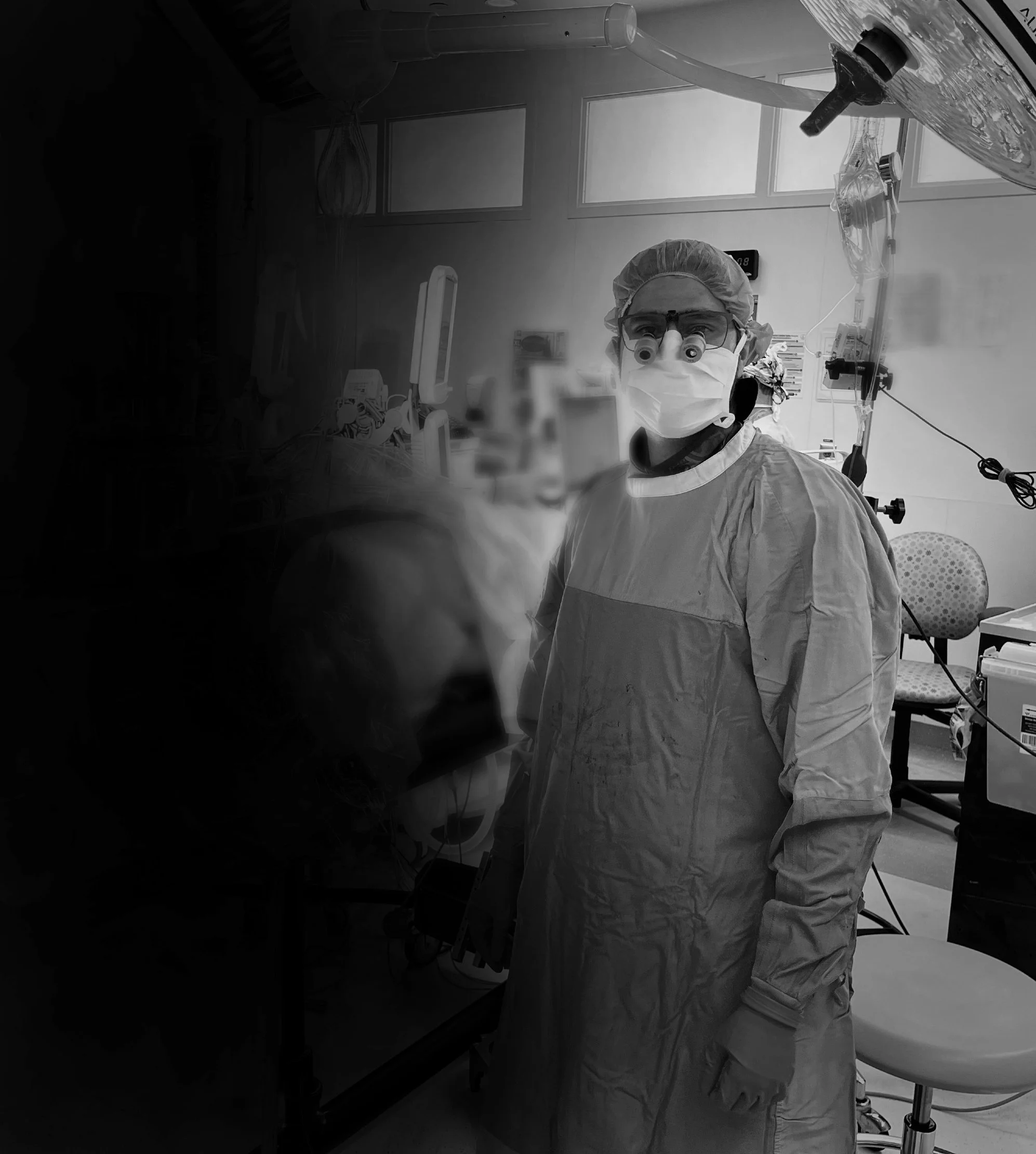 Dr. Parekh wearing a face mask and magnifying goggles in a clinical setting, with medical equipment and a window in the background.