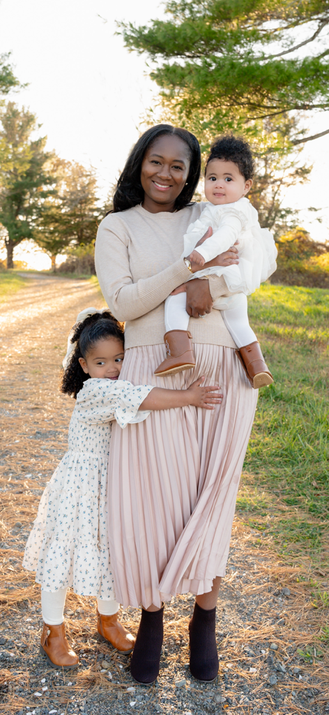 A smiling woman holding a young girl in white and brown boots, standing outdoors on a dirt path with trees and sunlight in the background.
