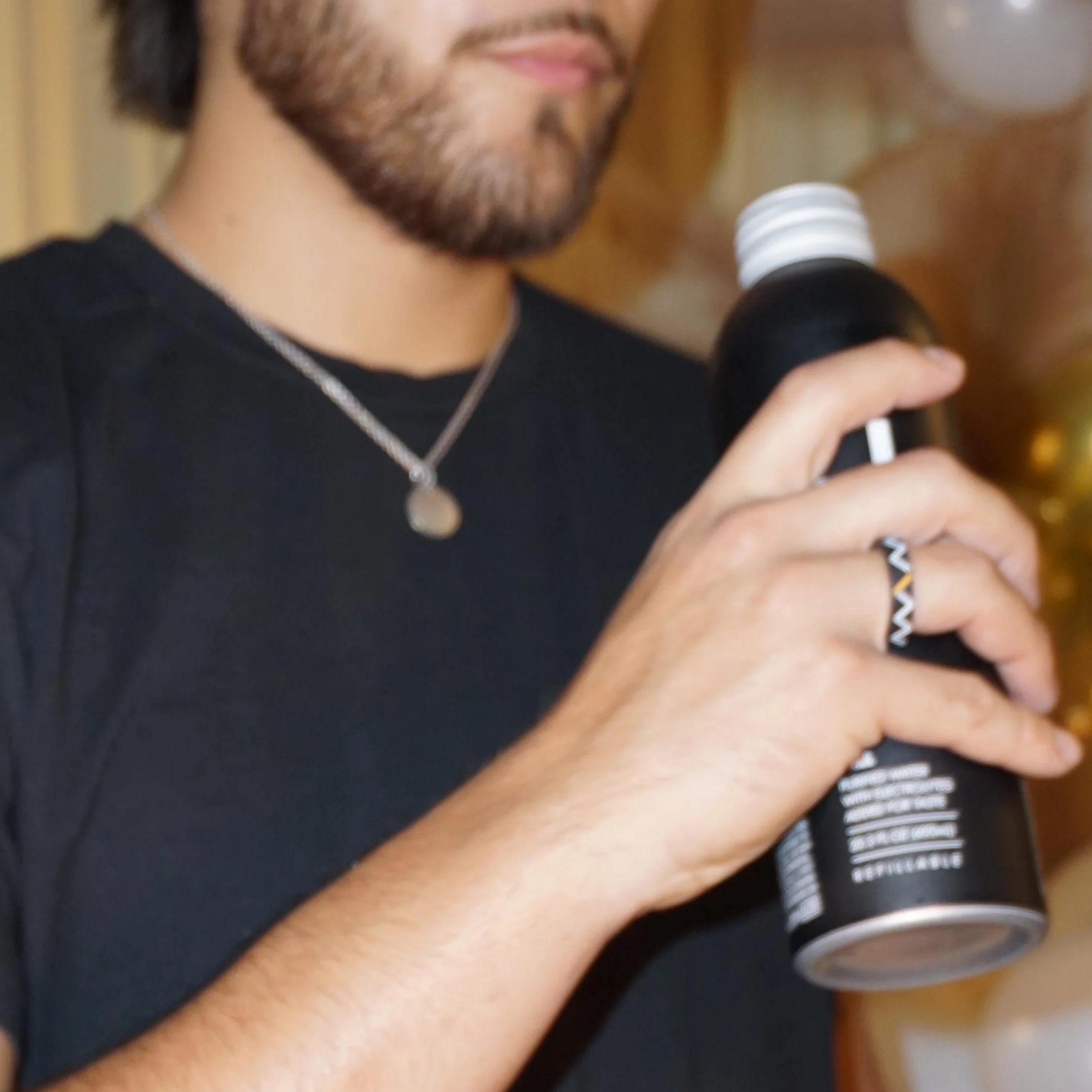A person with a beard holding a microphone and wearing a necklace, black shirt, and striped bracelet, with text visible on the shirt.