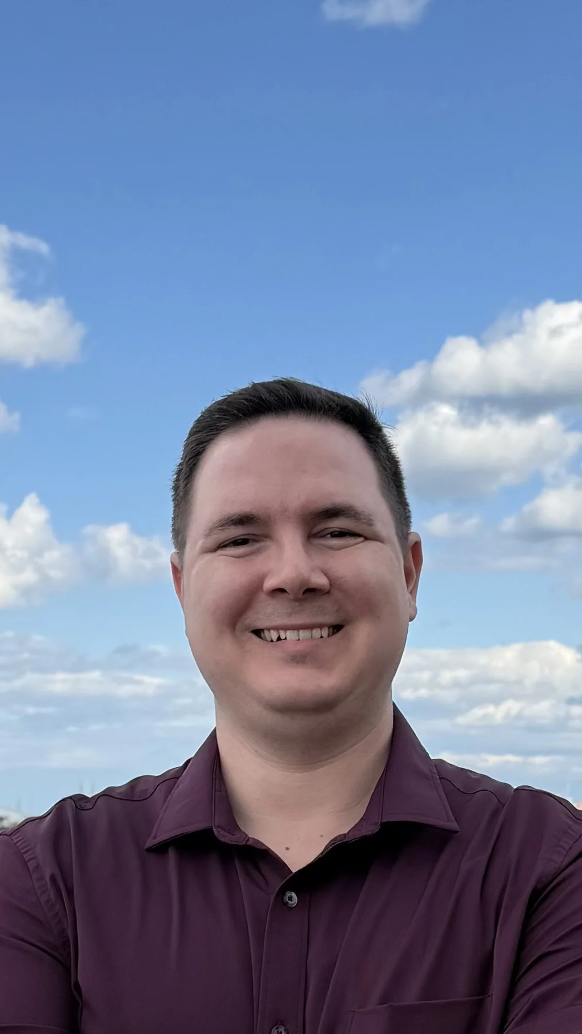 A man smiling outdoors with a blue sky and scattered clouds in the background.