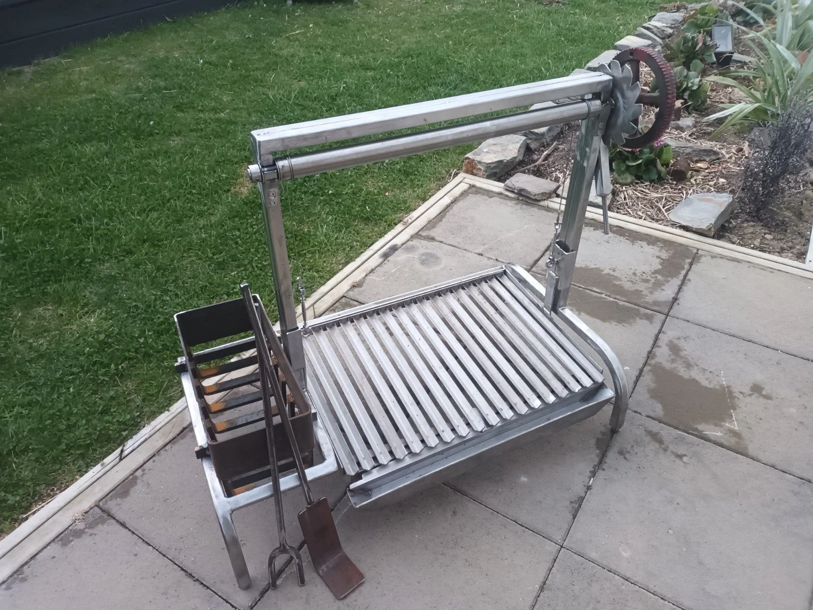 Outdoor metal grill with grilling tools hanging on side, placed on concrete patio with green grass and plants in the background.