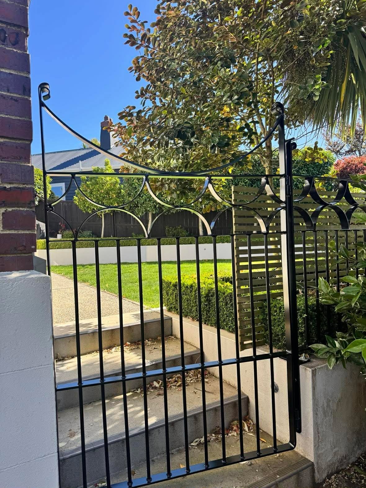 Black metal gate with decorative arches and support rods, attached to brick and concrete wall, leading to a backyard with steps, green lawn, various trees, and a wooden fence under a blue sky.