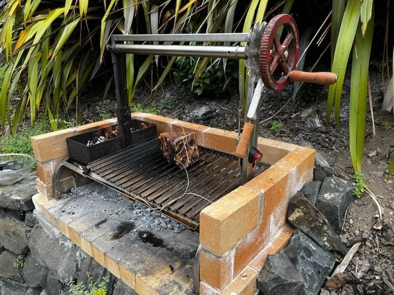 An outdoor barbecue grill with a rotisserie mechanism, a piece of meat on the spit, surrounded by rocks and greenery.