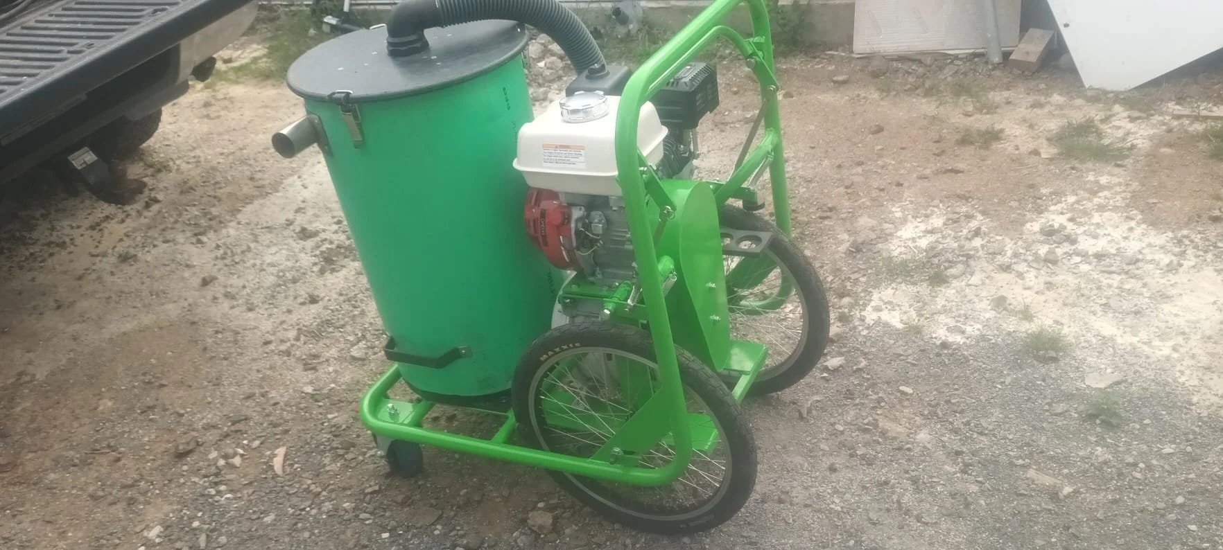 A green motorized street sweeper with a large waste container, mounted on a two-wheel frame, parked on a gravel surface outdoors.