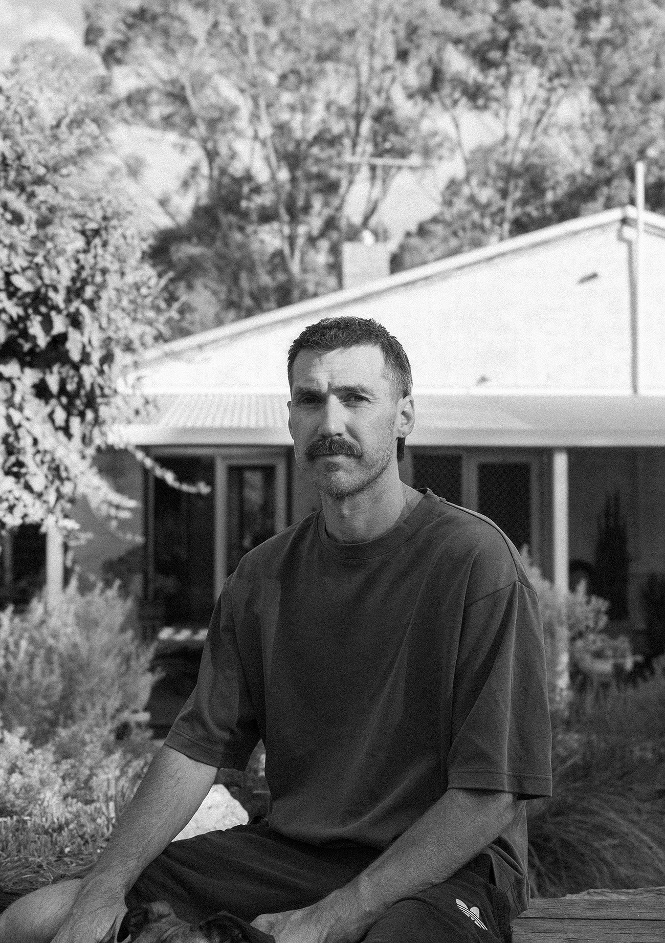 Black and white photo of a man sitting outdoors in front of a house and trees, wearing a dark T-shirt and shorts, with short hair and a mustache.