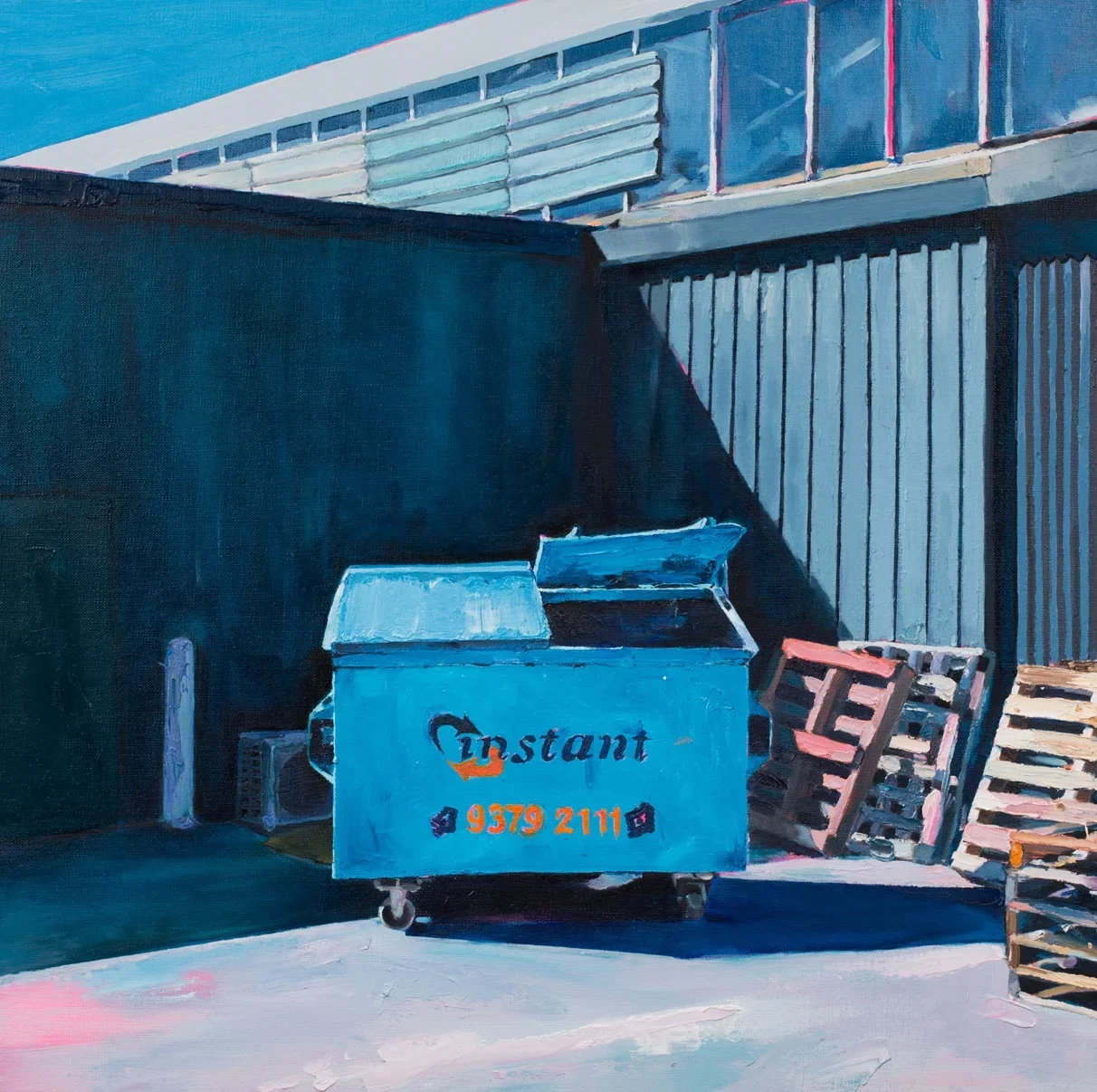 A blue dumpster with the word 'Instant' and a phone number, placed in an alleyway next to a dark wall, with wooden pallets stacked nearby and a corrugated metal fence in the background.
