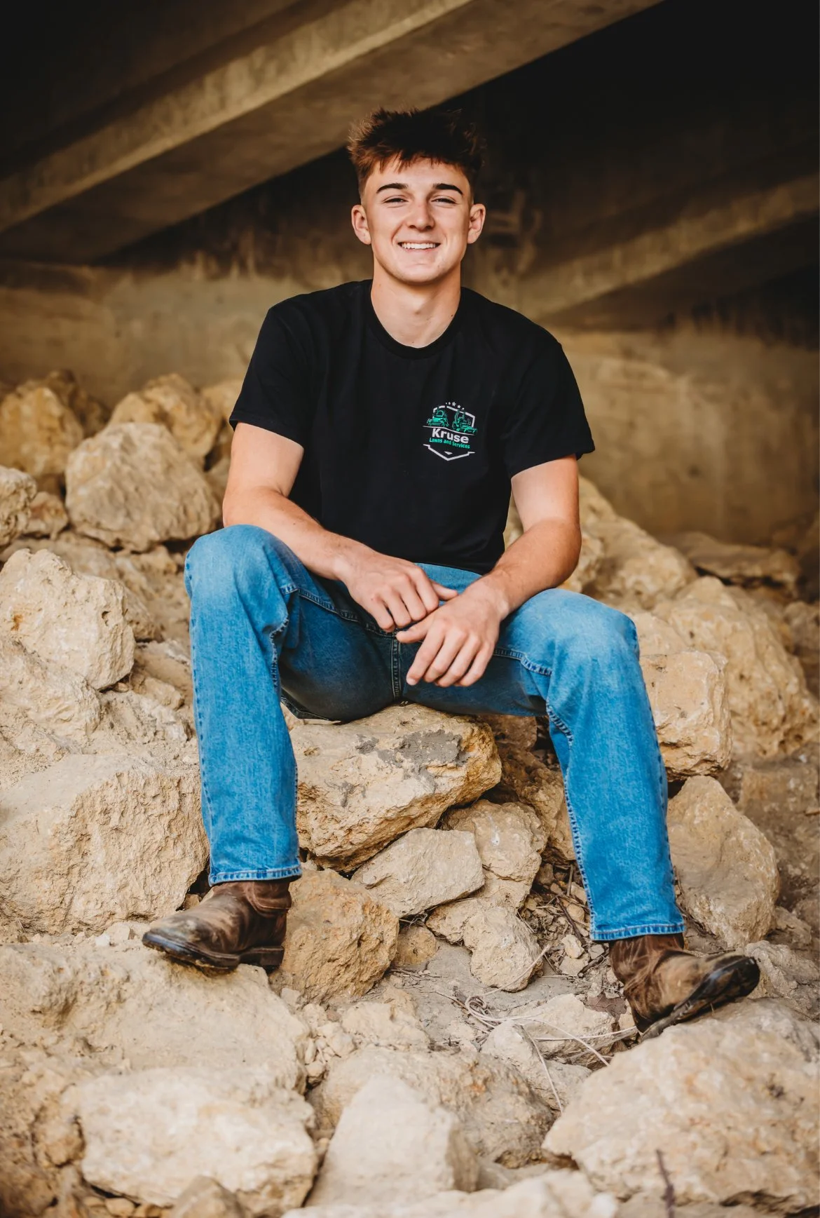 Young man sitting on rocks under a bridge, smiling, wearing black shirt, blue jeans, and work boots.