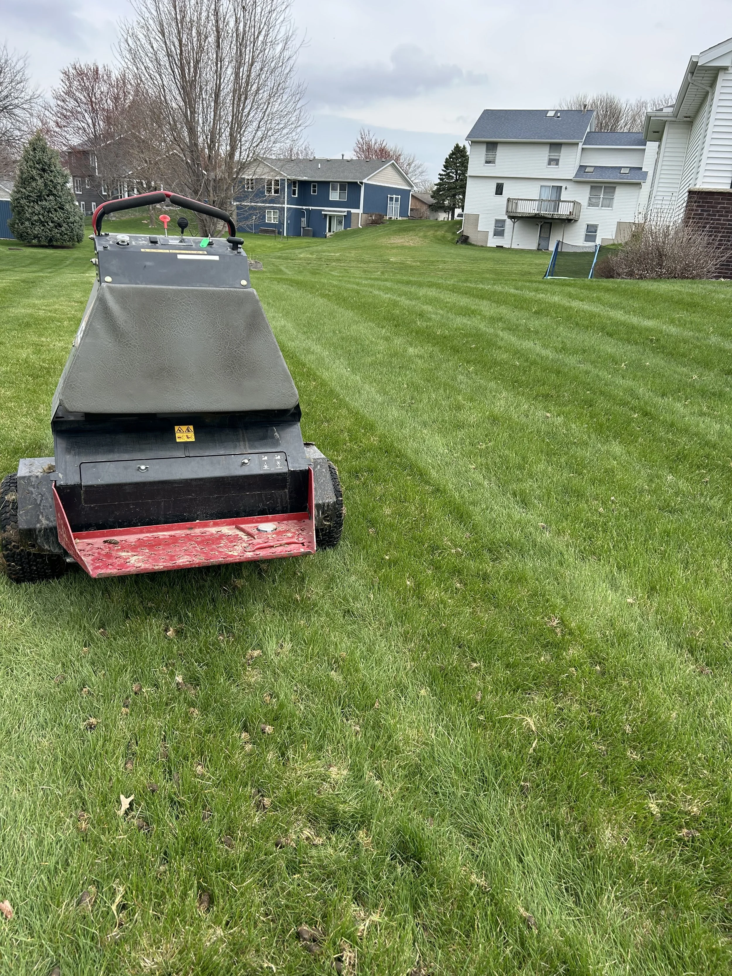 A lawn mower on a well-maintained grassy backyard with houses and trees in the background, cloudy sky overhead.