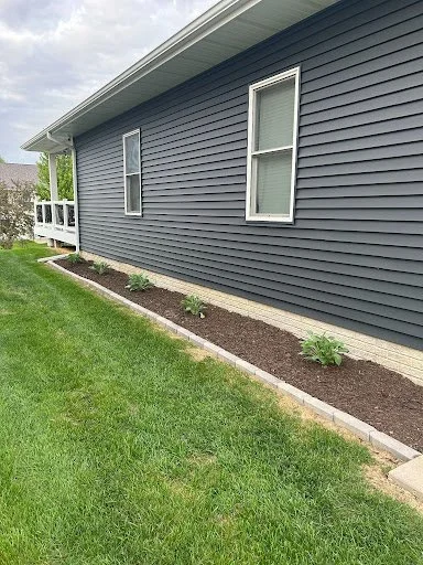 Side view of a house with blue siding, two windows, a small porch with railing, and a newly planted flower bed with small green plants along the foundation, next to a well-maintained lawn.