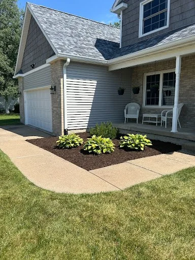 Front yard of a house with a concrete walkway, green grass, and a landscaped flower bed with plants, front porch with a bench, and a two-car garage.