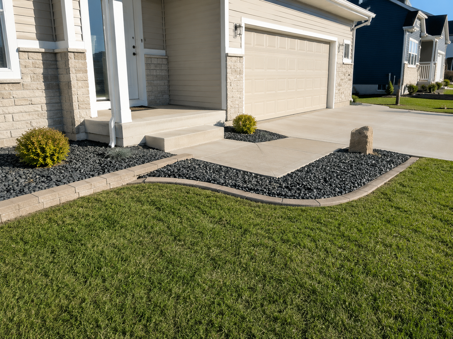 Front yard with a concrete sidewalk, two small plants, rocks, and a rock in front of a house with a beige garage door.