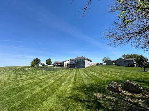 A wide grassy field with a house and trees in the background under a blue sky with some streaks of white clouds.