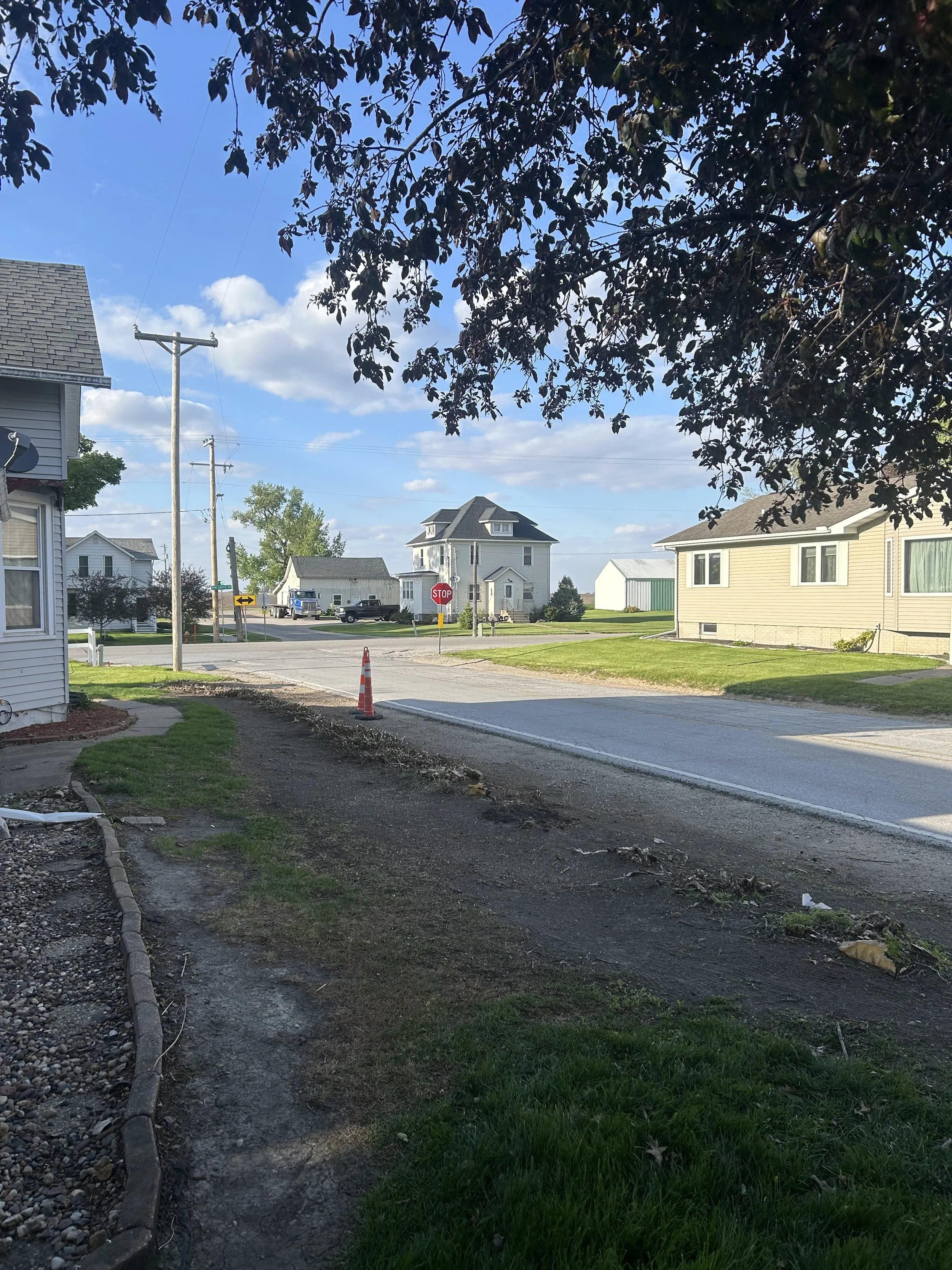 A suburban neighborhood scene showing a street with houses, a stop sign, utility poles, a sidewalk, and trees with a blue sky and scattered clouds overhead.