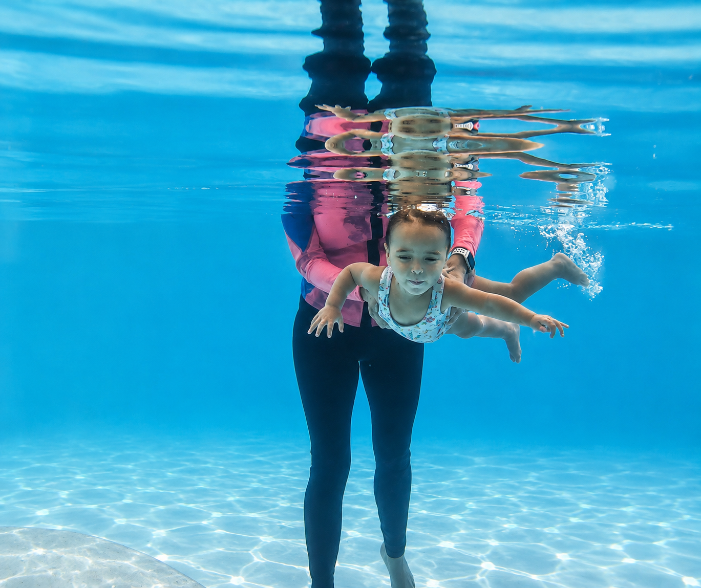 A young girl swimming underwater in a pool with an adult supporting her from below.