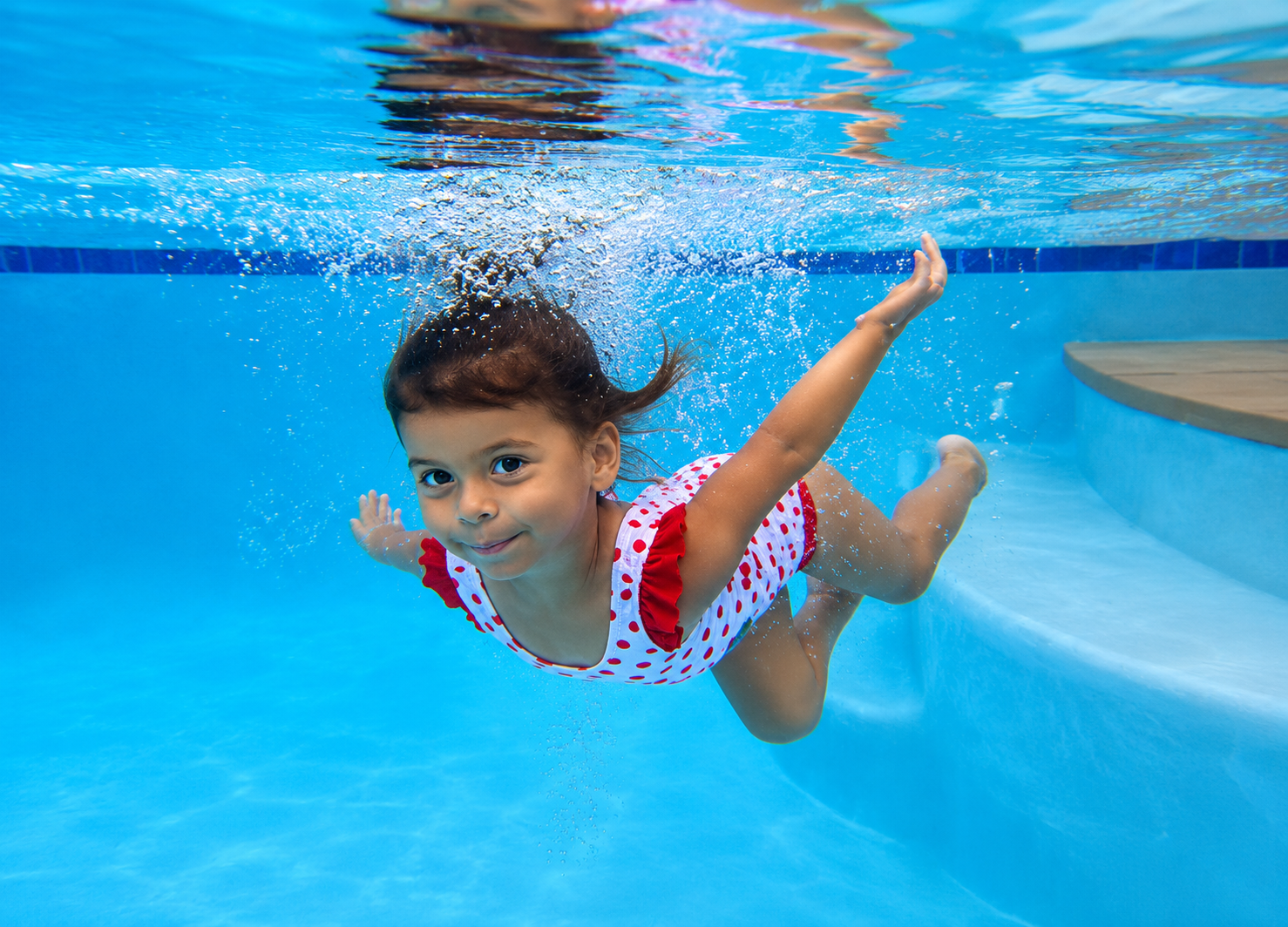 Young girl swimming underwater in a pool, wearing a white swimsuit with red polka dots and red ruffle details, smiling at the camera.