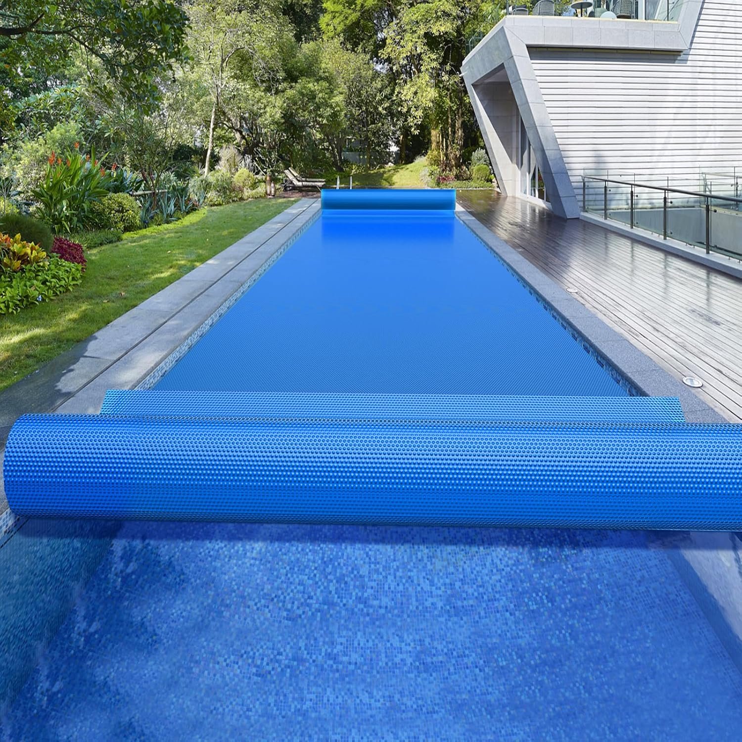 A modern outdoor swimming pool with a blue cover, surrounded by a wooden deck, lush green trees and plants, and a contemporary building with glass and concrete walls.