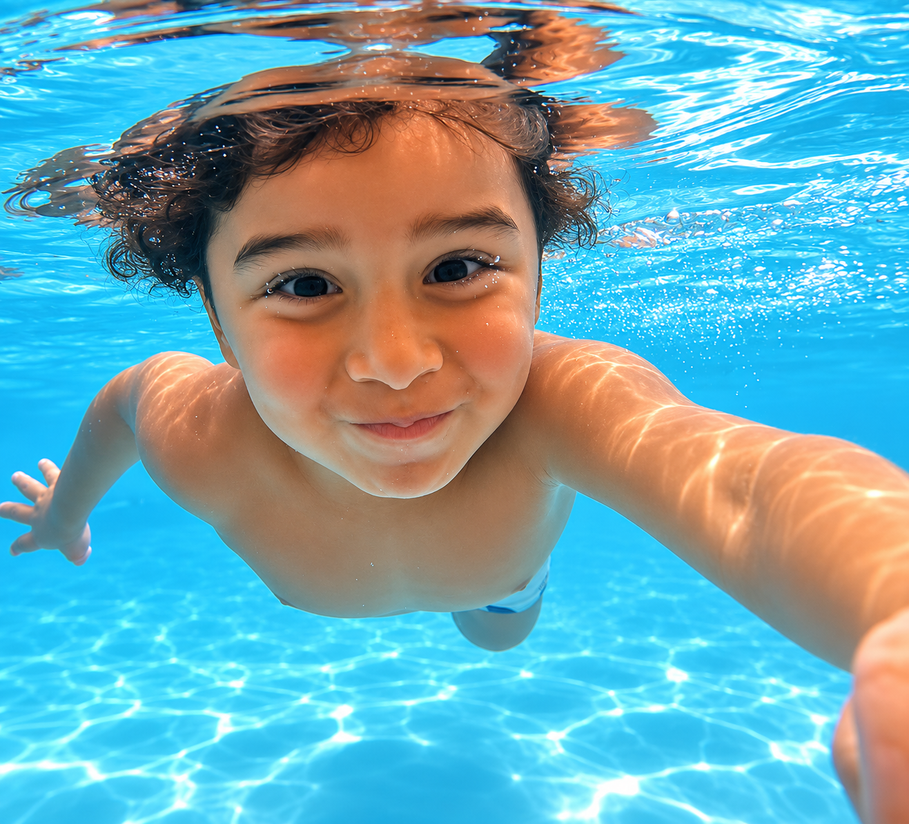 A young boy swimming underwater in a pool, smiling at the camera.