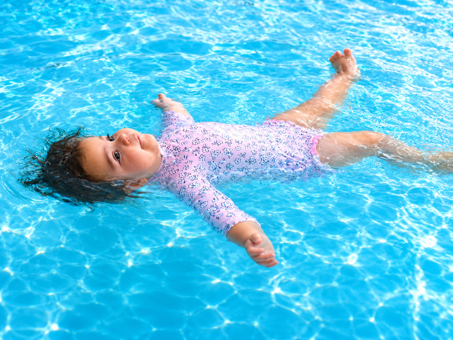A young girl floating on her back in a swimming pool, wearing a pink and purple floral swimsuit, with her arms outstretched and hair spreading in the water.