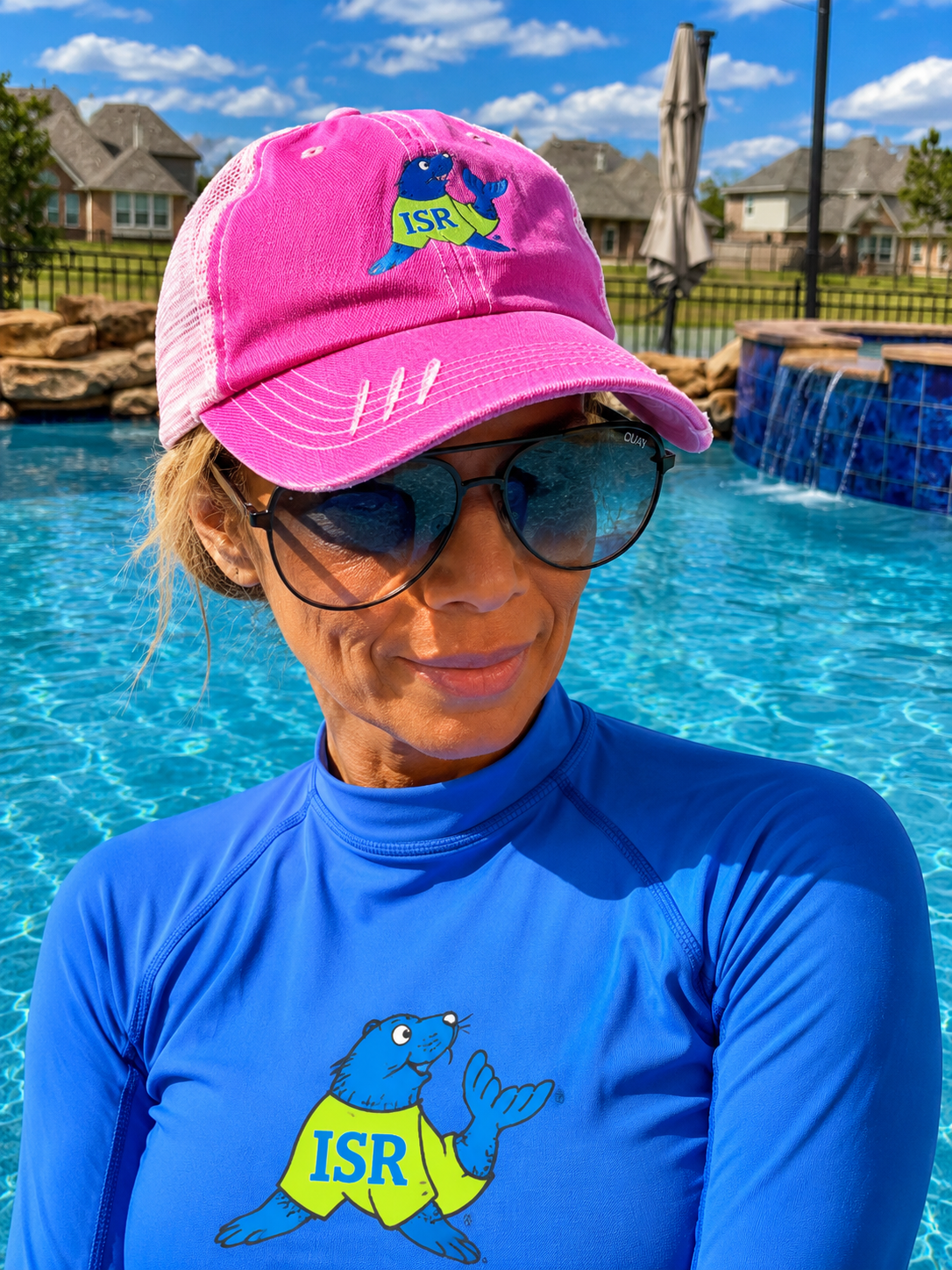 Woman wearing sunglasses, a pink cap with a cartoon otter and 'ISR' logo, and a blue rash guard with a cartoon otter and 'ISR' logo, standing poolside with water and houses in the background.